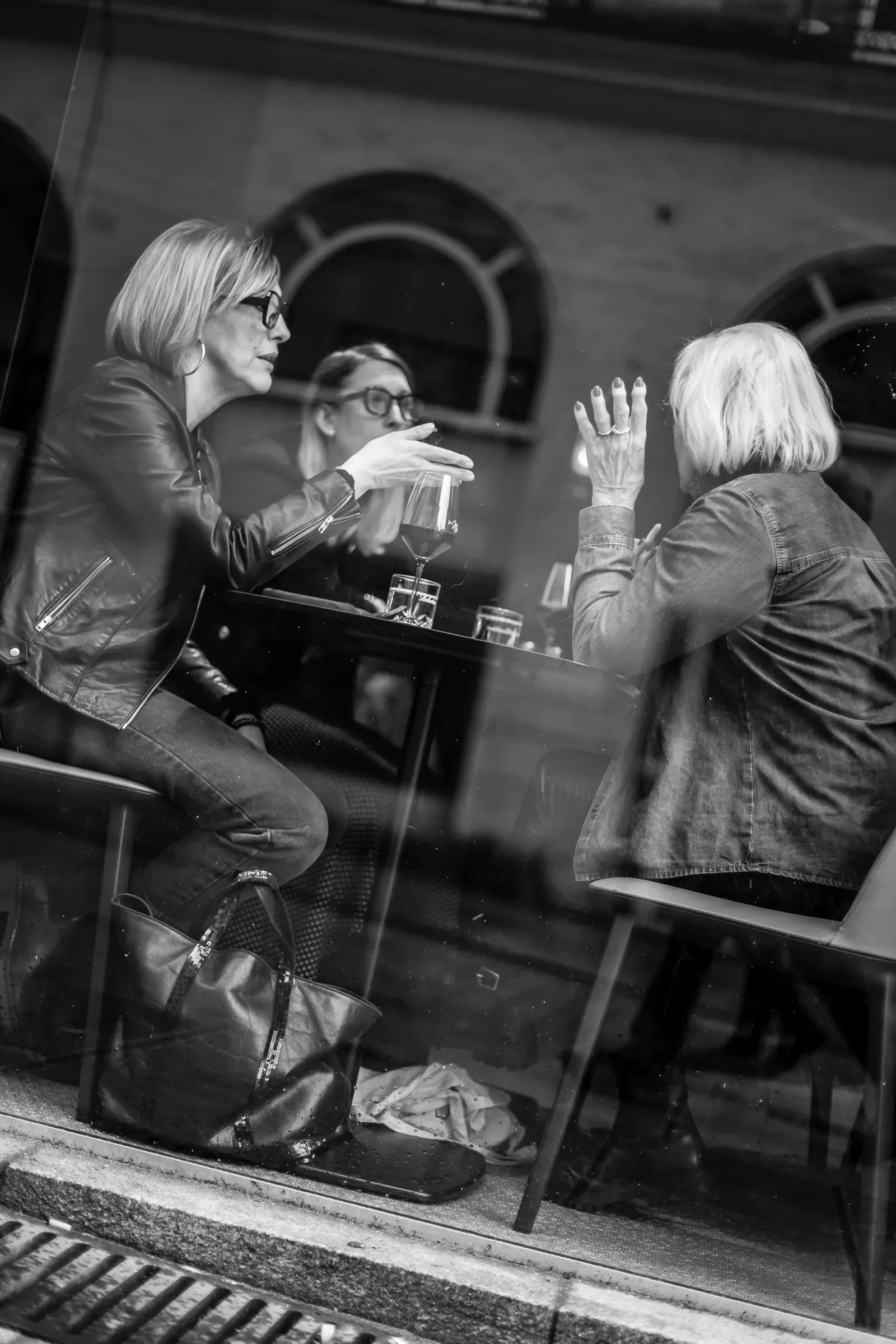 Three women seen through a café window, deep in conversation over glasses of wine