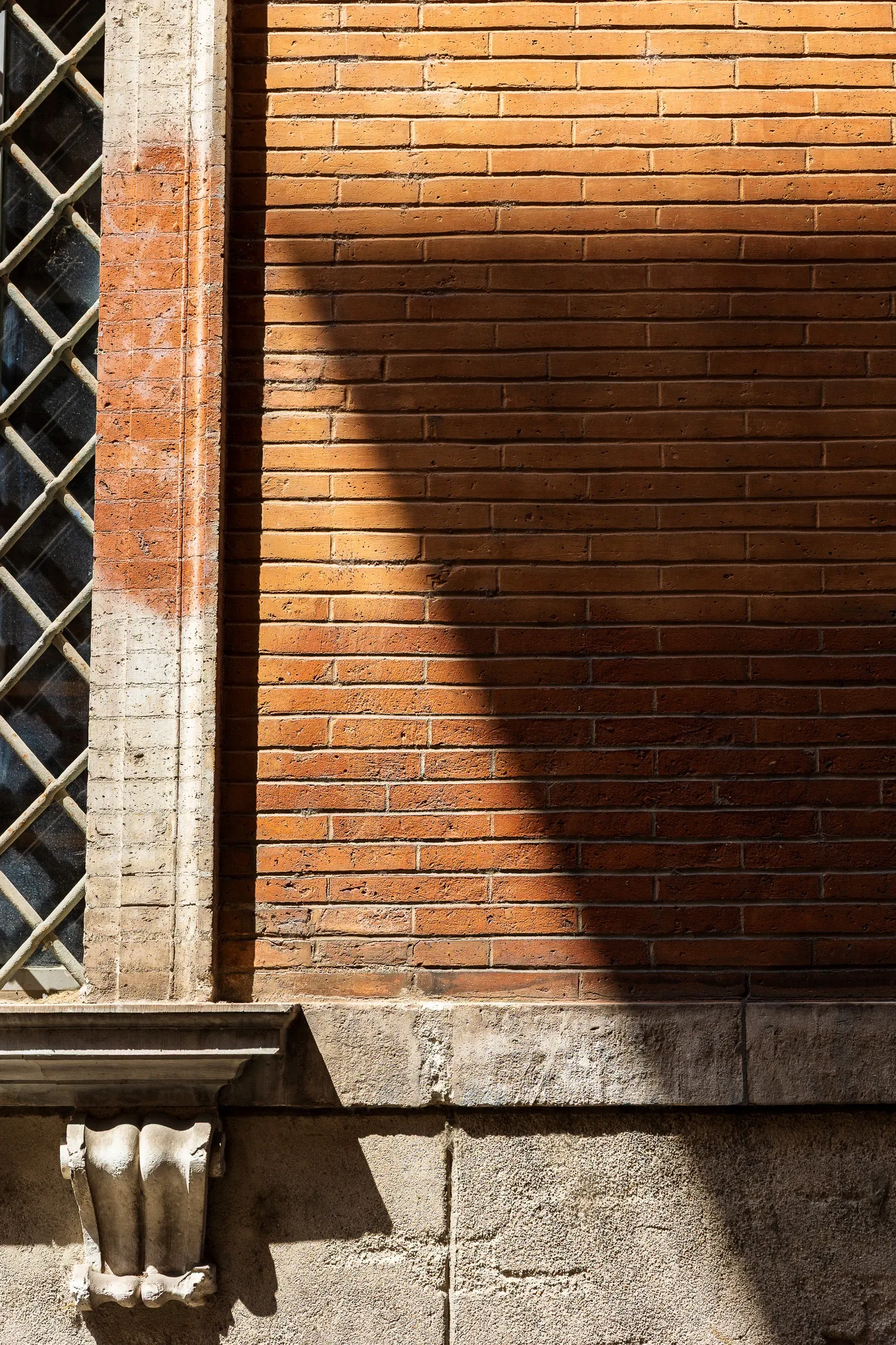Diagonal shadow cutting across a terracotta brick wall, stone column and iron window grille on the left side of the frame, Toulouse