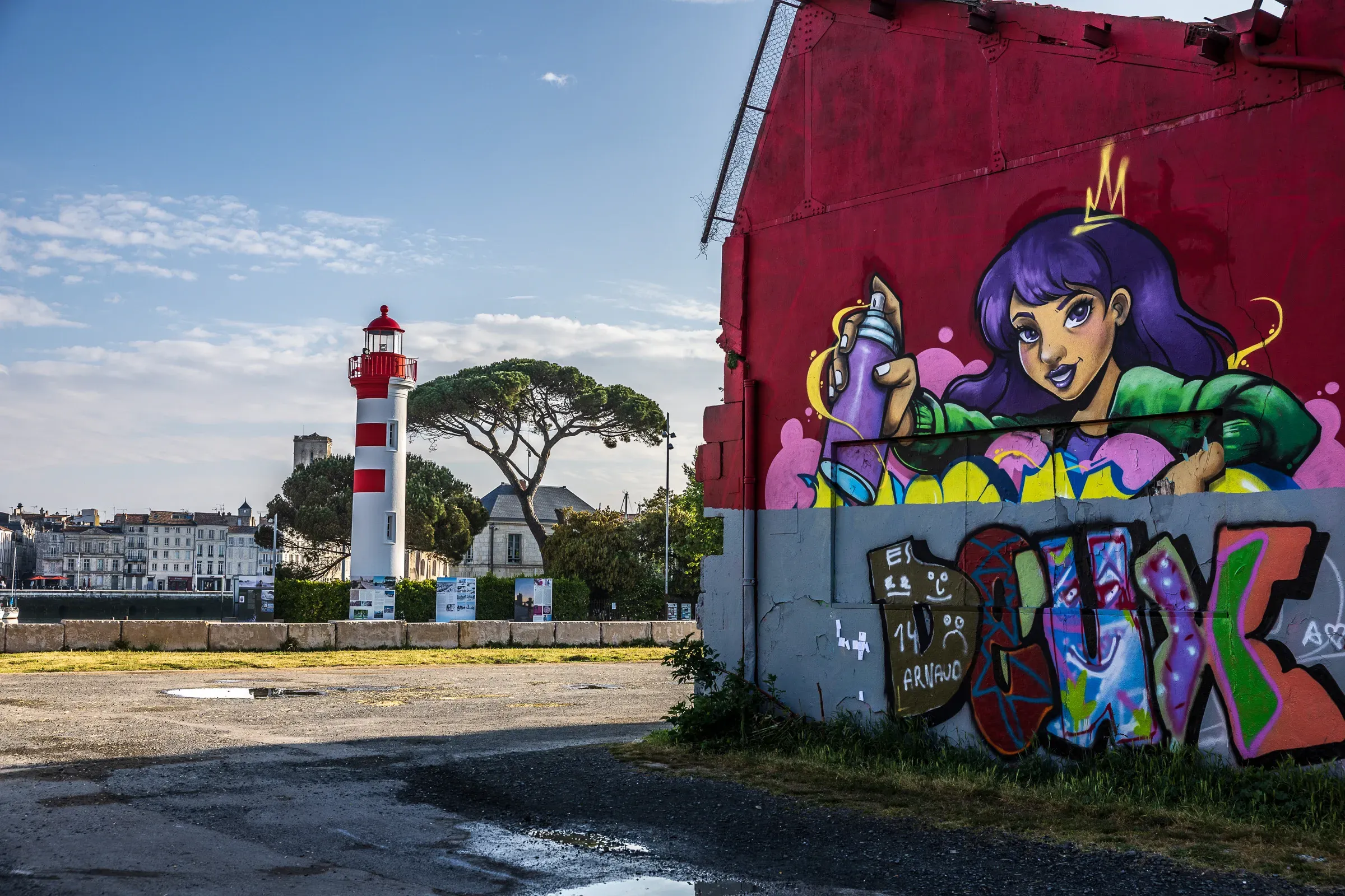 La Rochelle's red-and-white lighthouse seen across a port yard, next to a large mural of a crowned woman with purple hair holding a spray can, colorful tags below