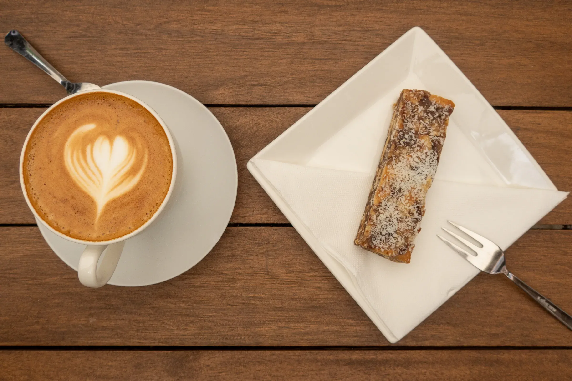 Cappuccino with a latte-art heart beside a pastry on a white napkin, wooden cafe table