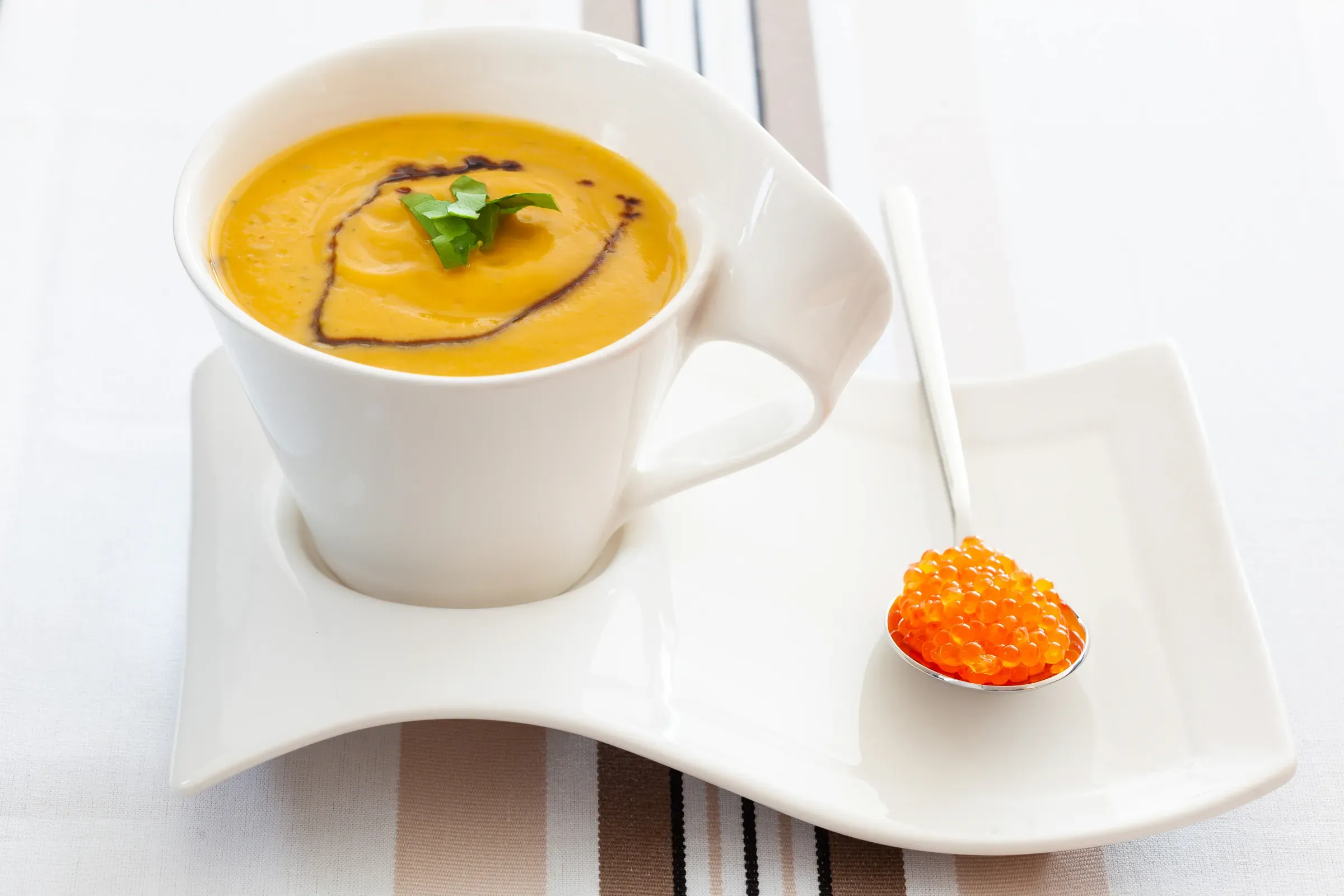 Squash velouté in a white mug with a herb swirl on top, small spoon of salmon roe resting on the saucer beside it, striped linen background