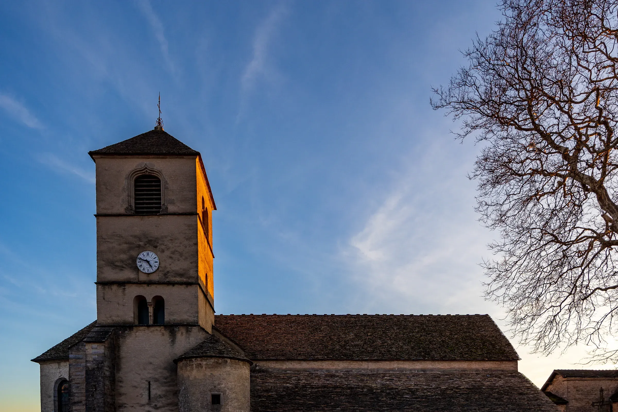 Romanesque church tower with a clock face bathed in warm golden sunrise light against a blue sky, bare tree to the right