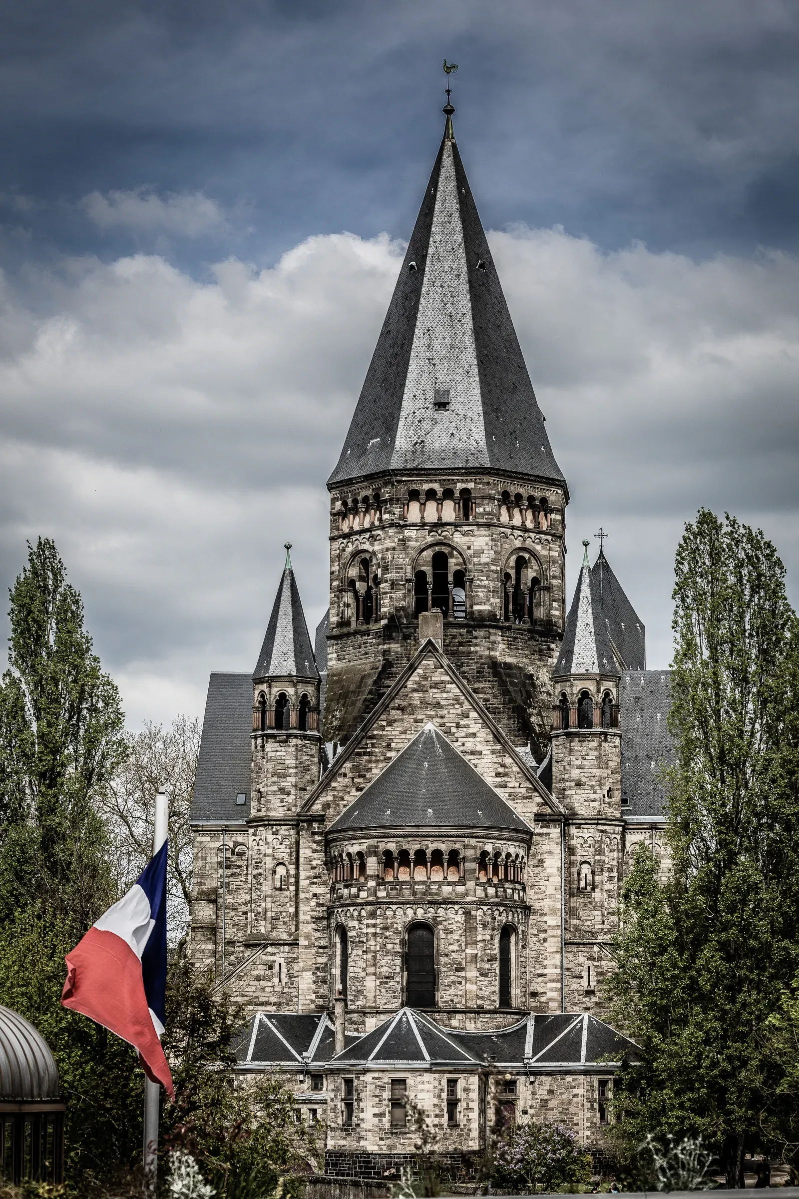 Temple Neuf seen from the front with a French flag flying, dark storm clouds building, trees flanking the neo-Romanesque facade