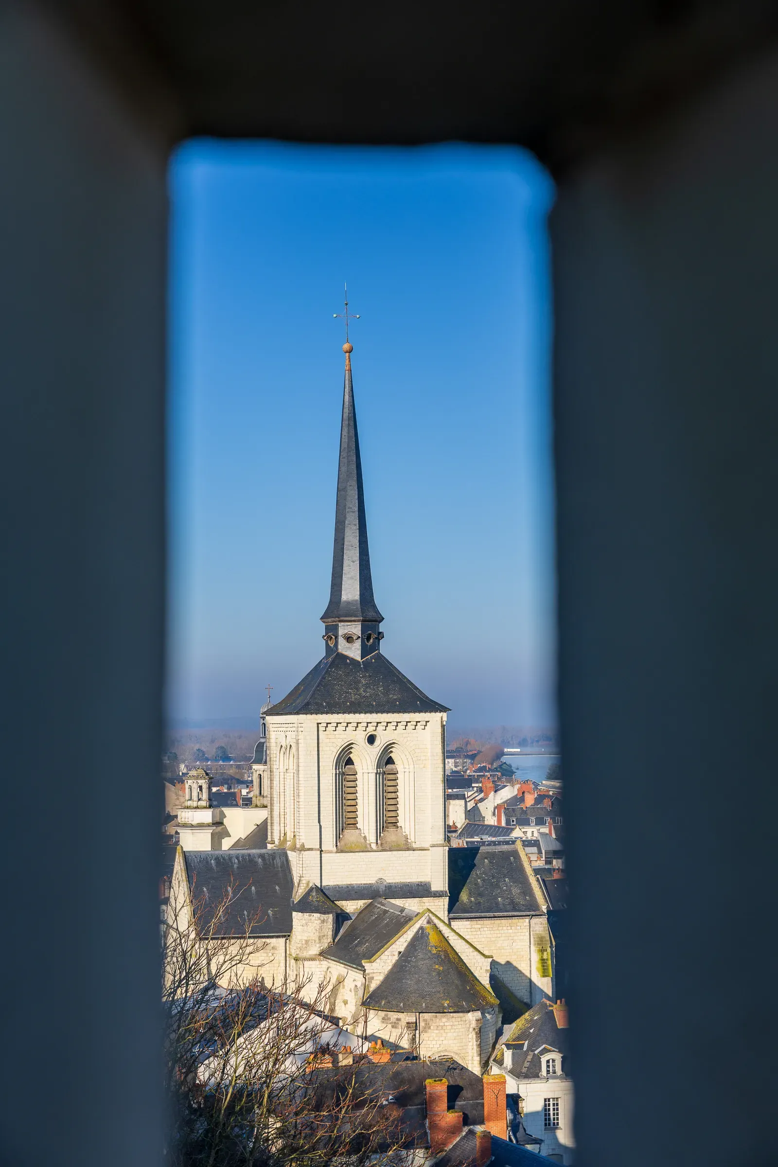 Church steeple framed through a narrow stone embrasure in the château wall, Saumur rooftops and river valley below in winter sun
