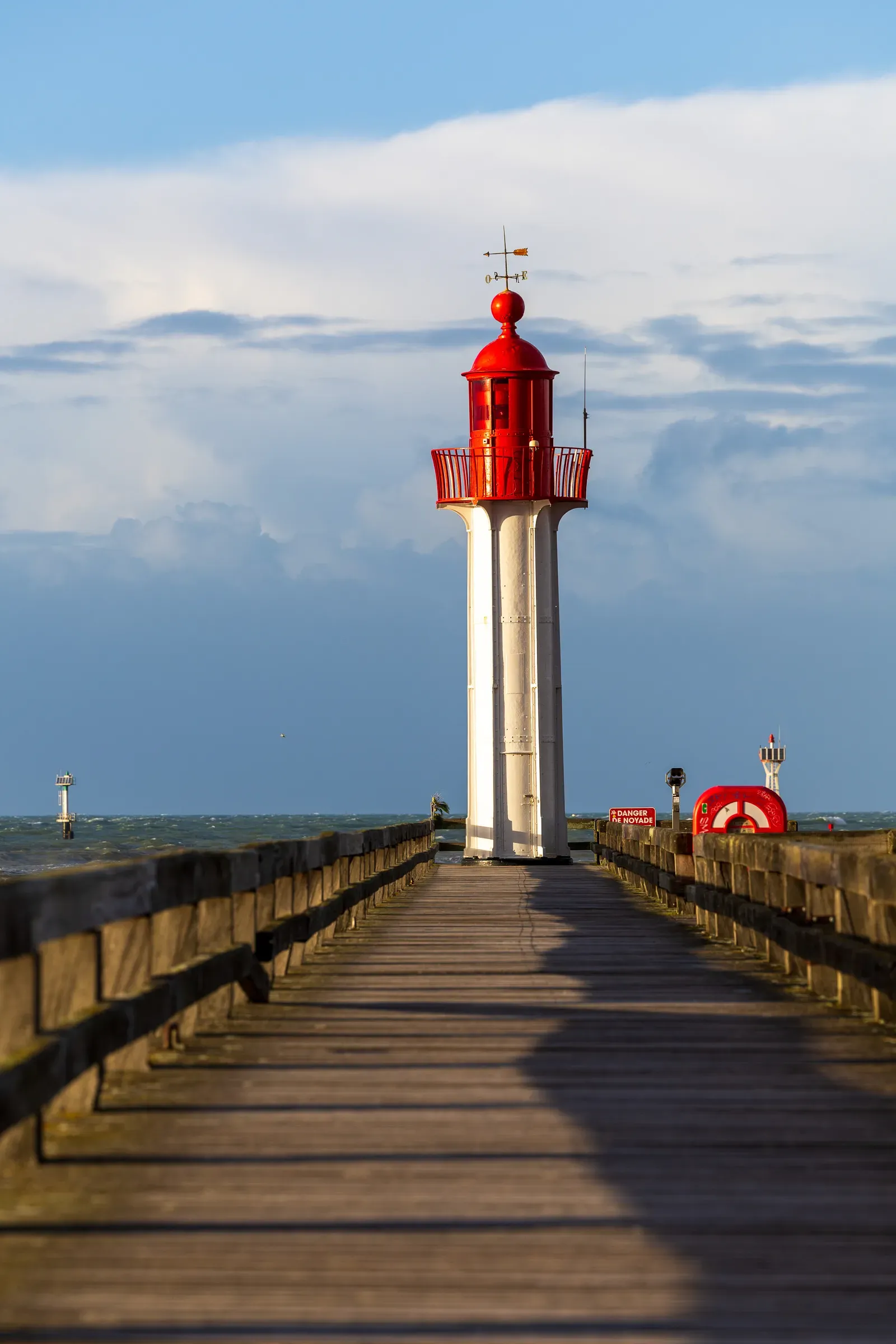 Red and white Trouville lighthouse at the end of a wooden pier seen from ground level, warm afternoon light, choppy sea on both sides, Trouville