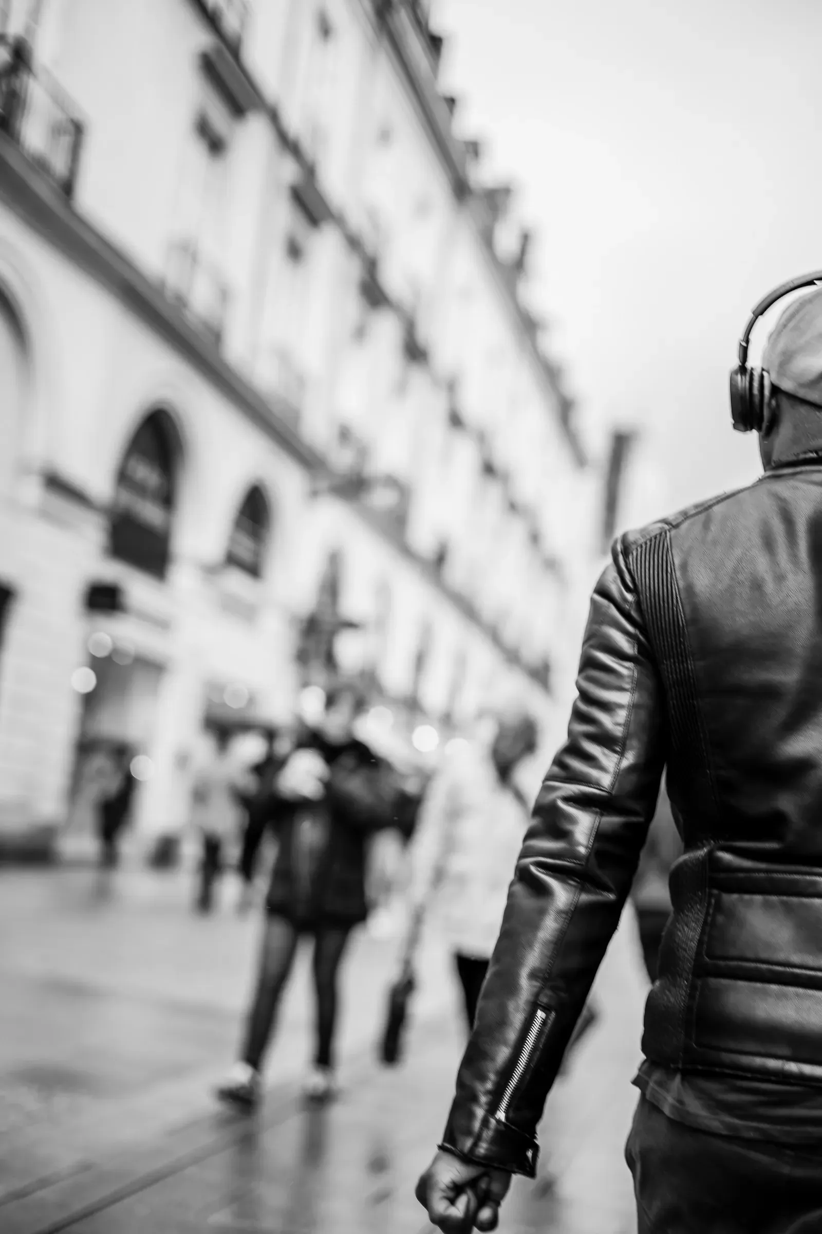 A man in a leather jacket, headphones and cap walks away down a street, pedestrians blurred ahead