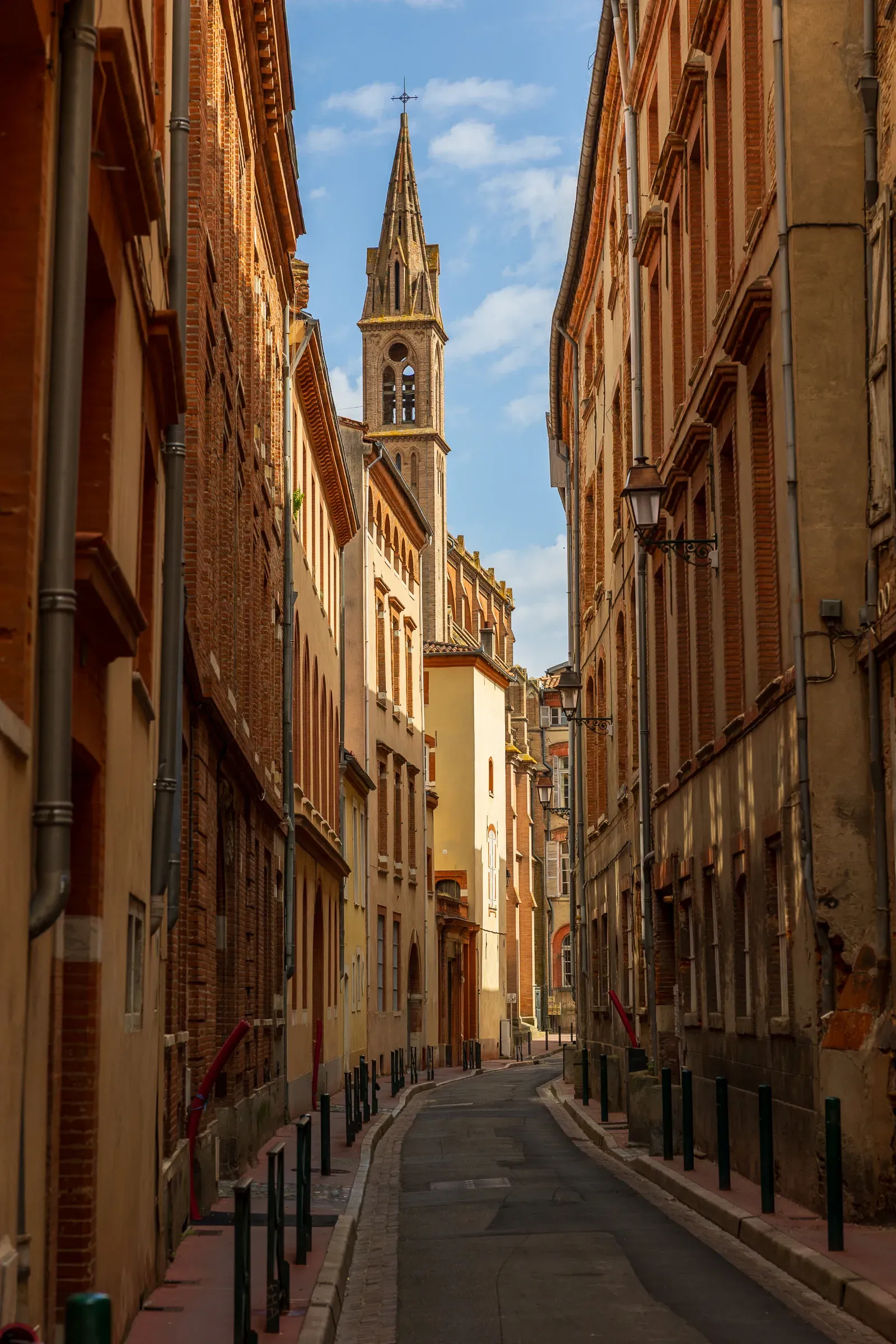 Narrow Toulouse street lined with pink and ochre brick buildings, a slender church steeple framed at the far end, Toulouse
