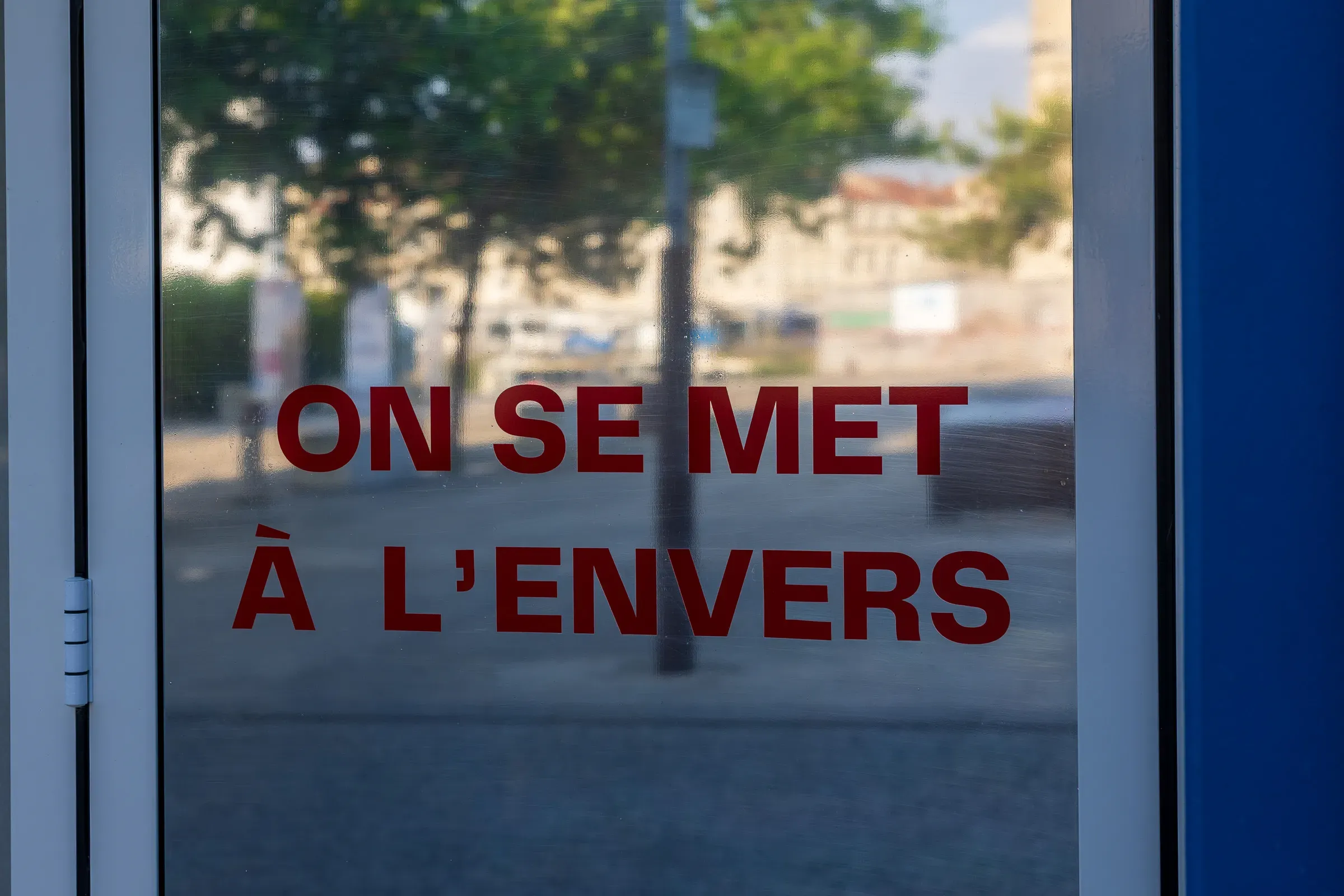 Glass door with bold red text reading 'On se met à l'envers', street and trees reflected in the glass behind