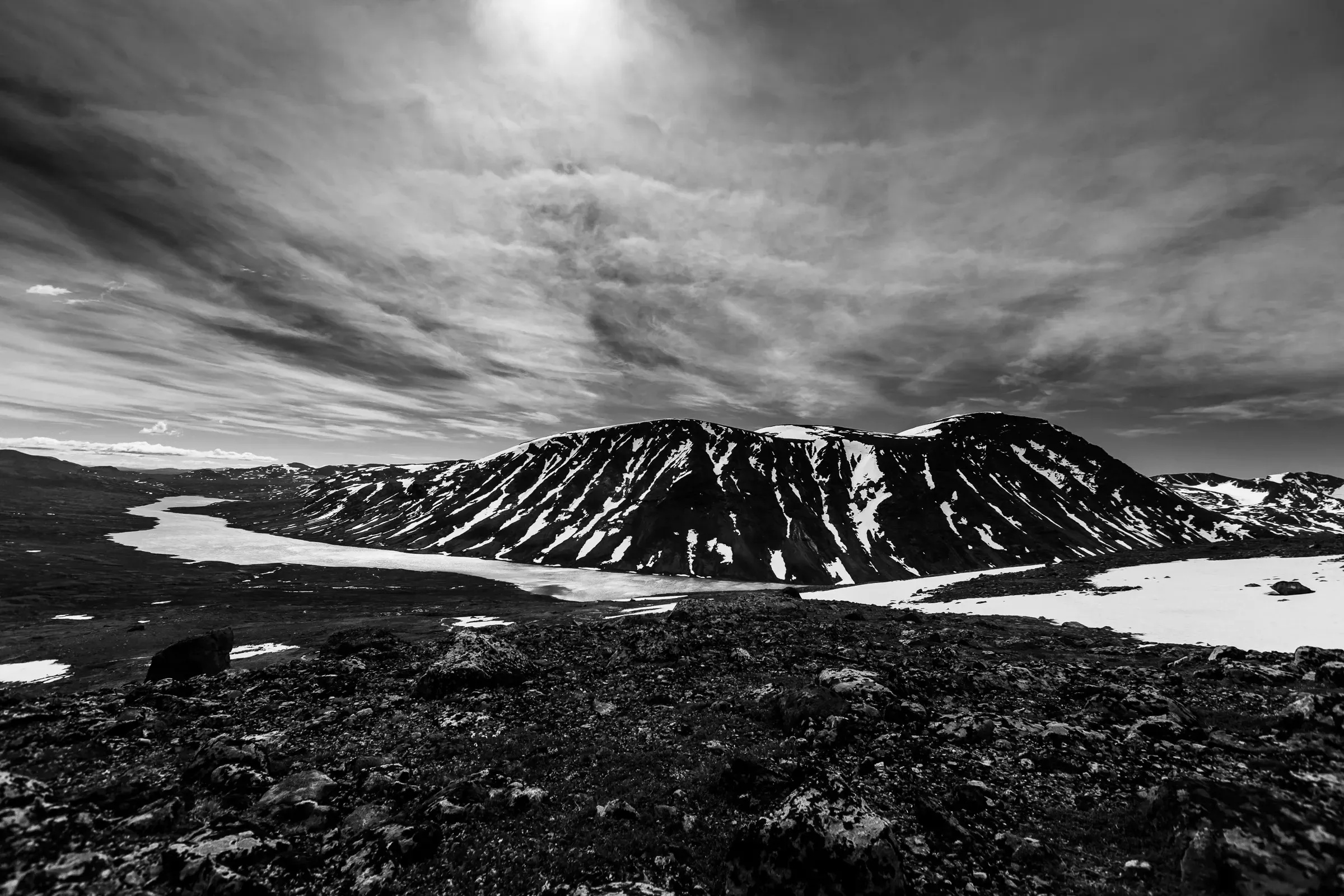 Snow-streaked mountain rising above a frozen valley lake on a Norwegian plateau, the sun burning through high cloud, volcanic rock in the foreground