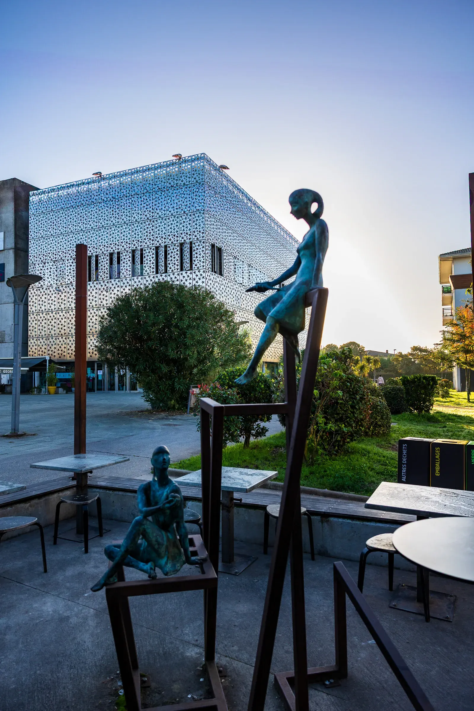 Two bronze sculptures of seated figures on metal frames in front of a perforated white cube building at dusk, Bordeaux