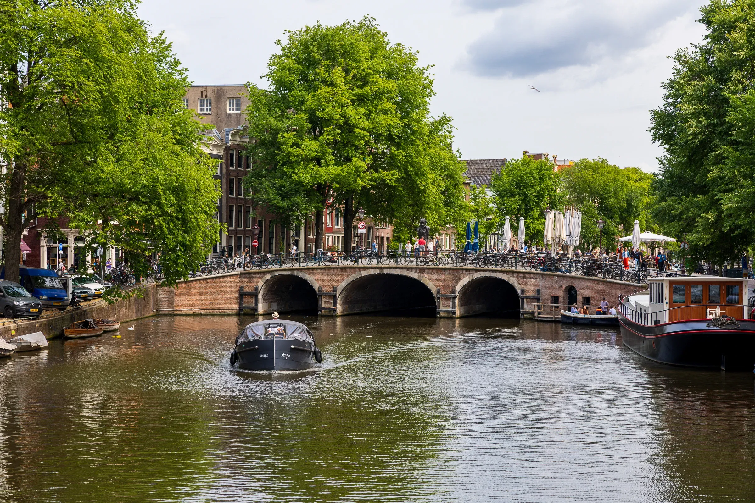 Dark barge passing under a brick arch bridge on a tree-lined Amsterdam canal, terrace diners on the right