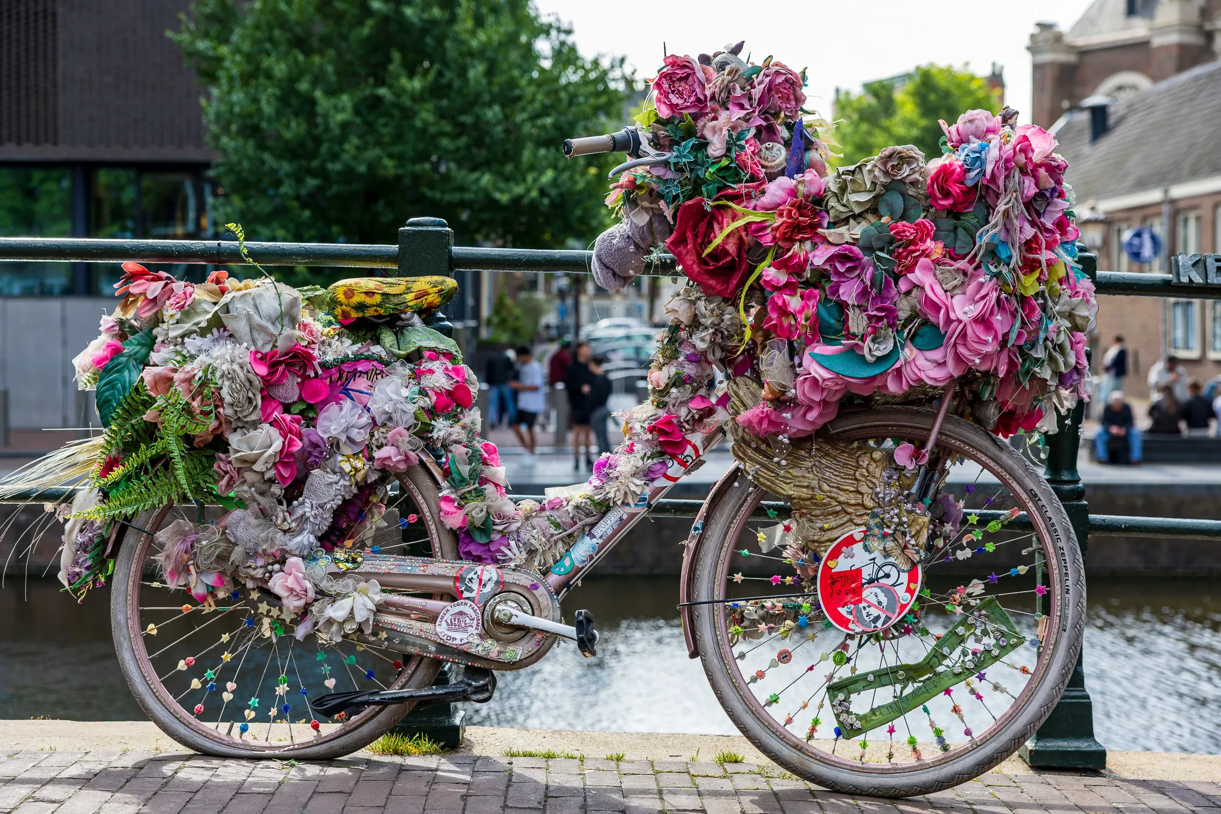 A bike buried under an explosion of pink and multicolored artificial flowers, locked to an Amsterdam canal bridge