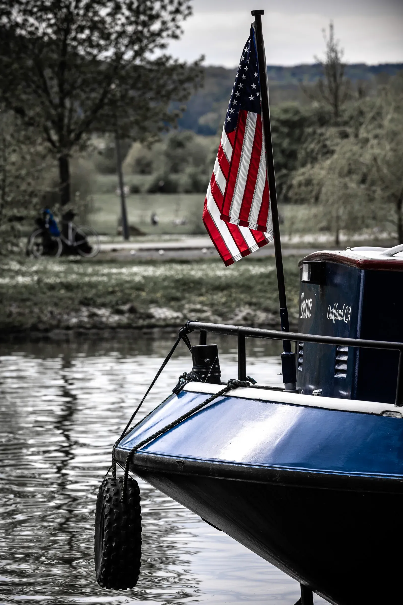 Bow of a blue boat from Oakland, CA flying an American flag, moored on the Moselle, selective color against a grey landscape