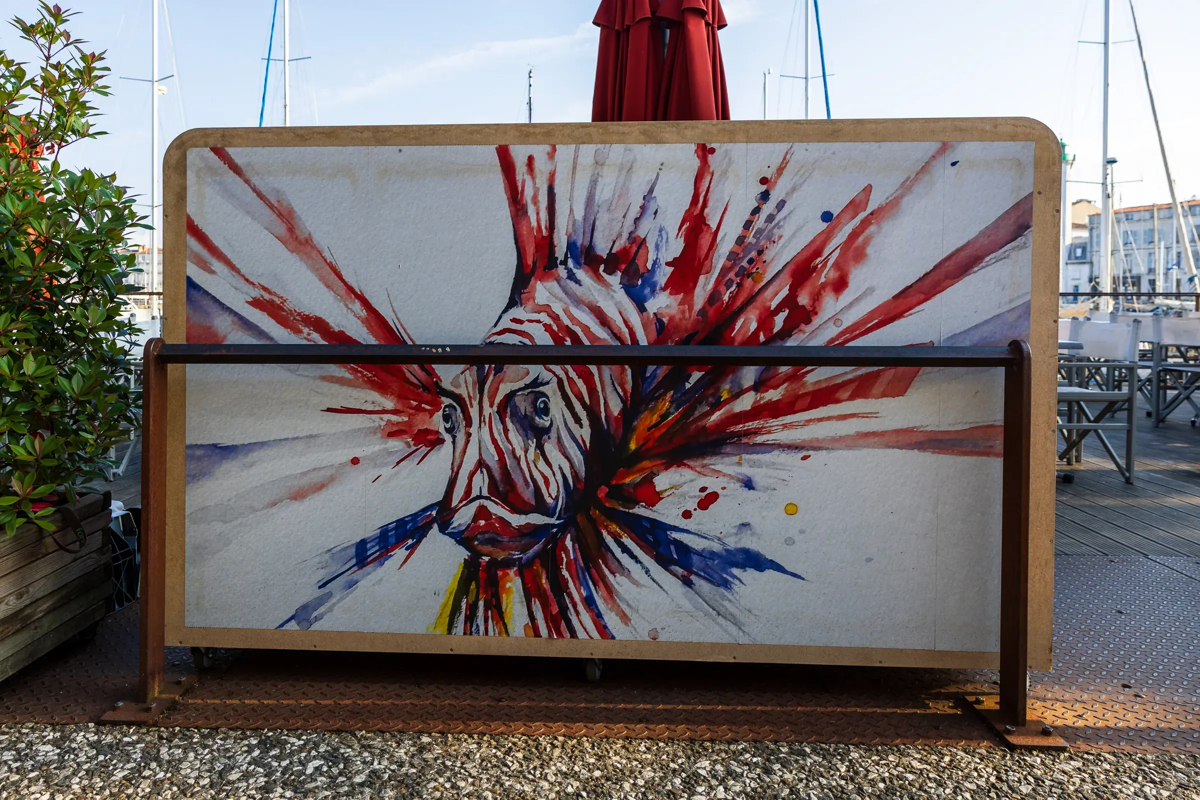 Outdoor artwork on a rusted metal stand — a rooster's head rendered in explosive red, blue, and yellow brushstrokes, moored boats and a furled umbrella visible behind