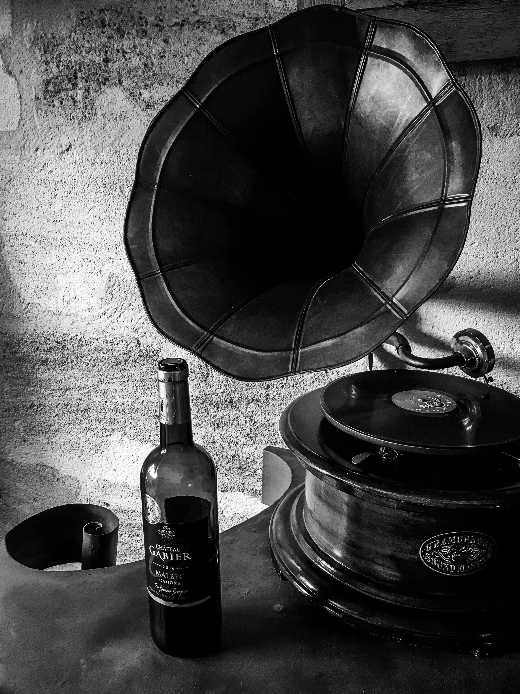 Antique gramophone with a large flared horn beside a bottle of Château Mabier Malbec, rough stone wall in the background