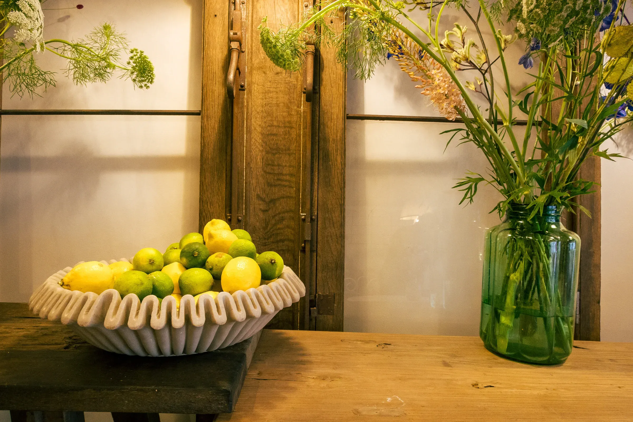 Ceramic bowl of citrus fruit and a green glass vase of wildflowers on a wooden table, rustic shuttered door behind