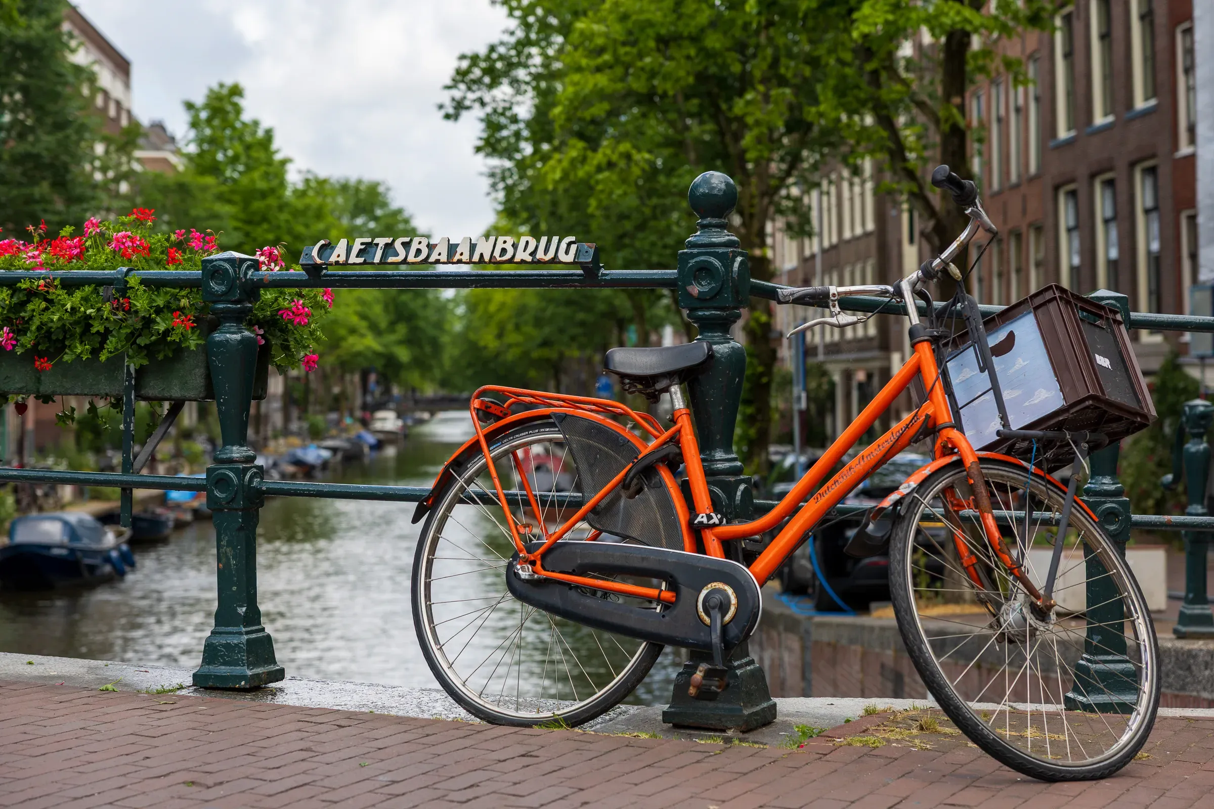 Bright orange Dutch bike with a wicker basket locked to the ornate Caetsbaanbrug bridge railing, canal behind