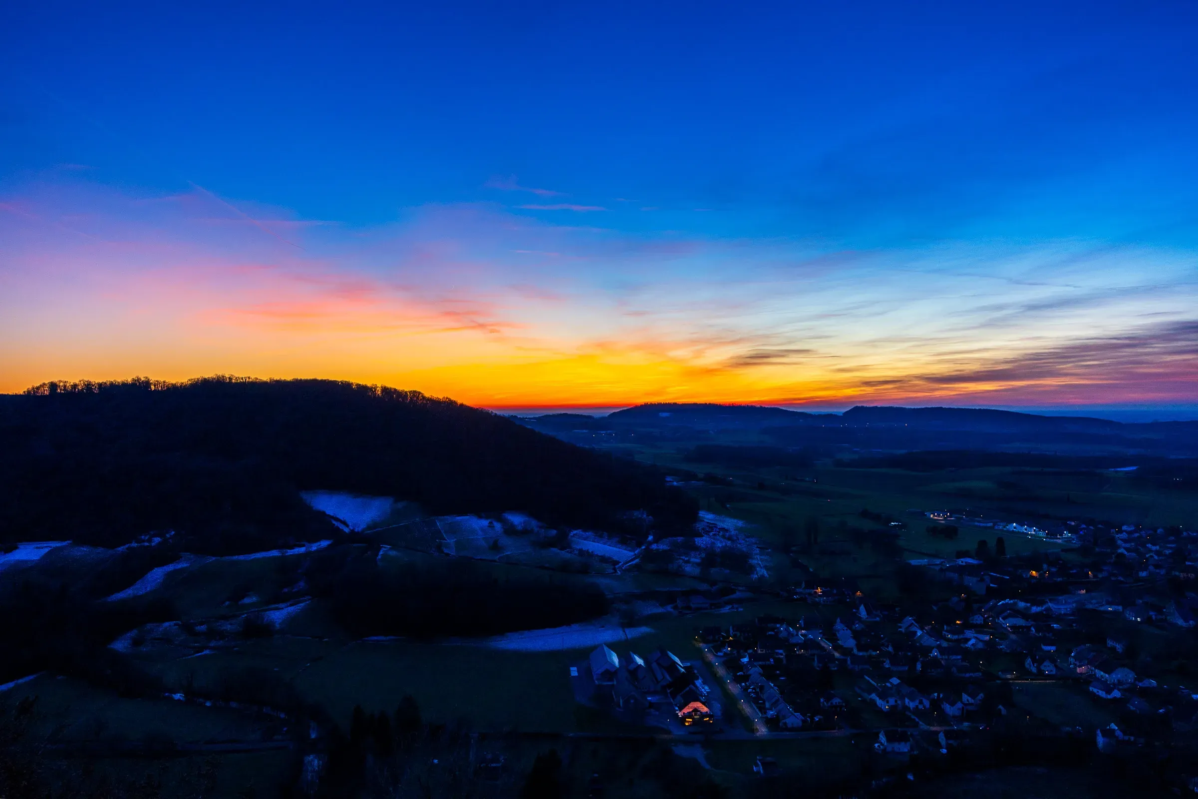 Aerial view of a Jura valley village at deep blue twilight, a streak of orange-yellow on the horizon, light snow on the hills