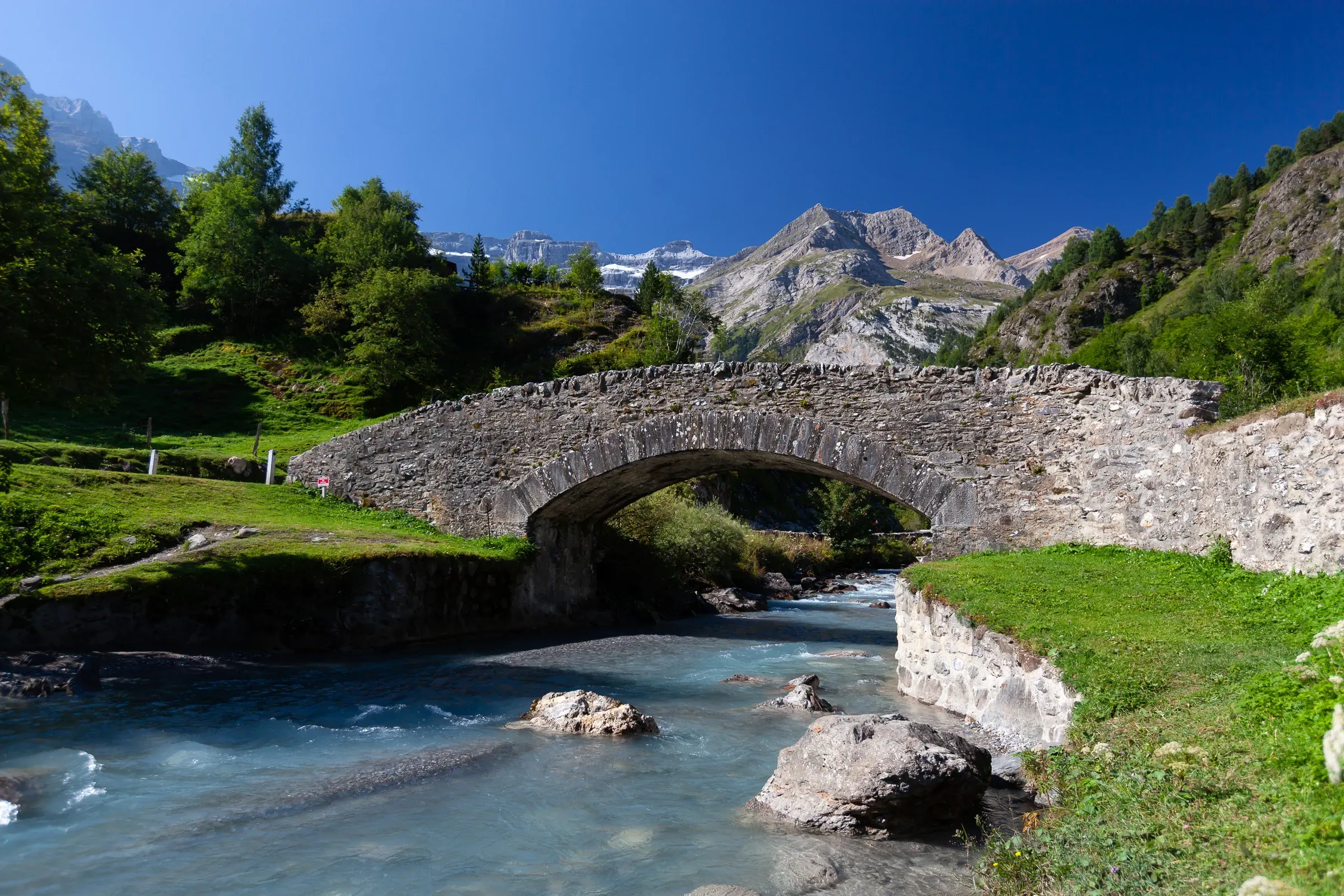 Stone arch bridge over a glacial turquoise river, Pyrenees — green alpine meadows and rocky peaks in the background