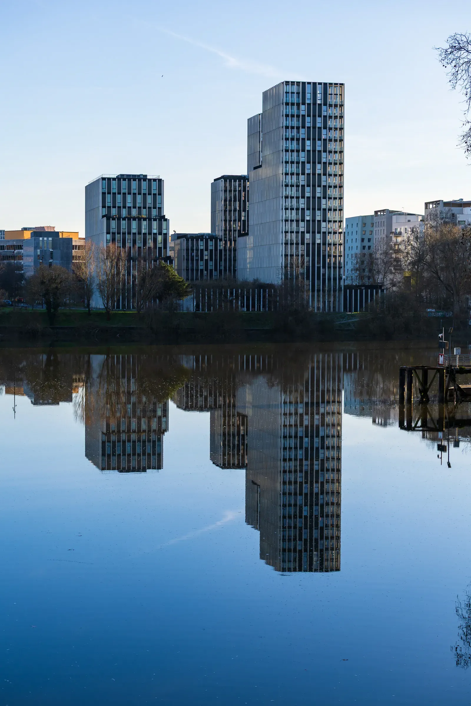 Cluster of modern glass and steel tower blocks reflected in a still body of water at the edge of a park, Nantes