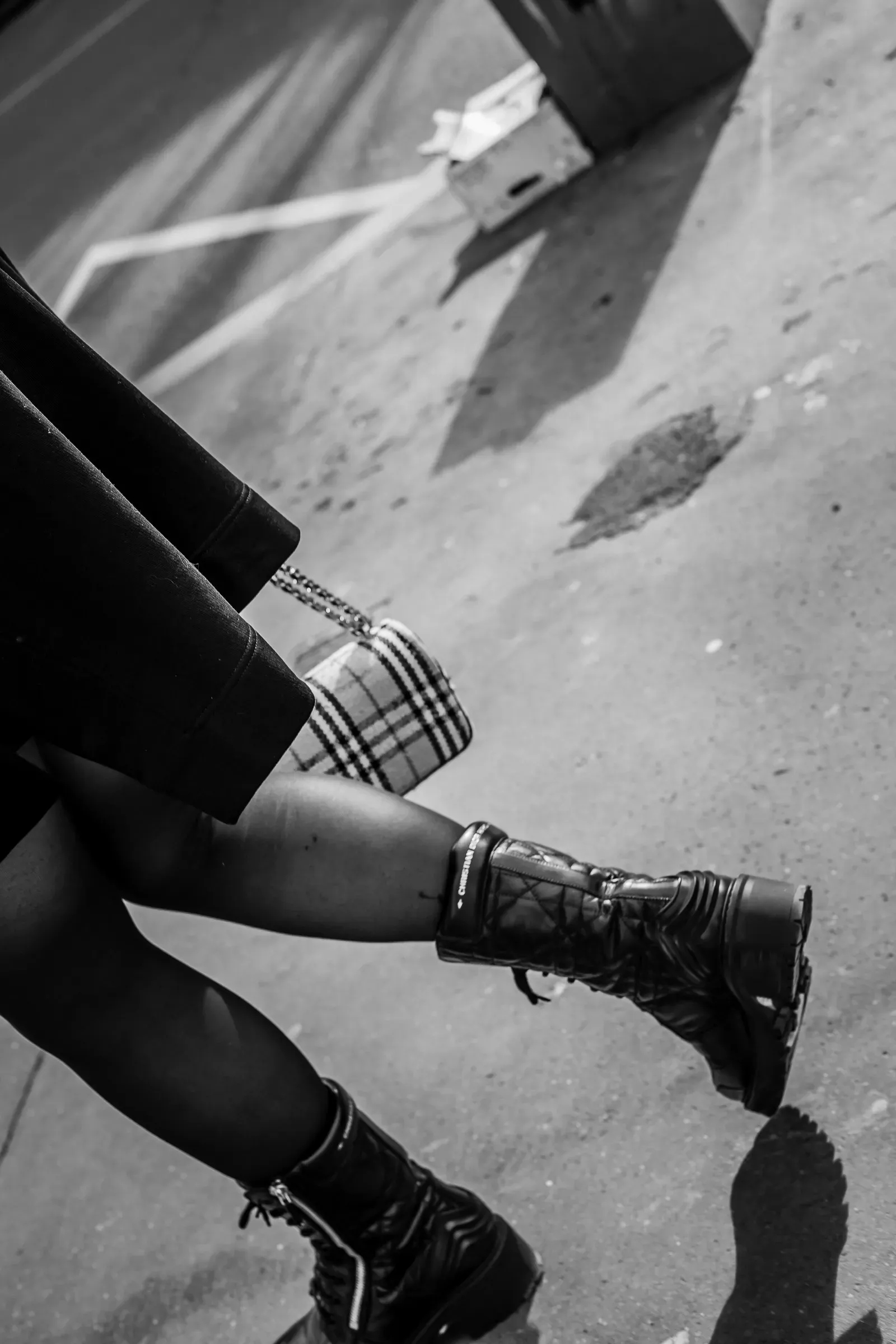 A tilted close-up of a woman's legs and boots crossing a street, her shadow cast diagonally across the pavement