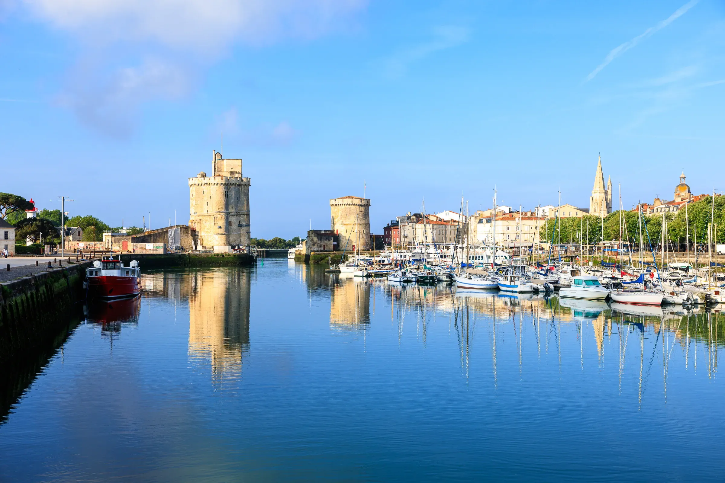 La Rochelle's medieval harbour at golden hour, Tour Saint-Nicolas and Tour de la Chaîne reflected in still water, sailboats moored to the right, church spire beyond