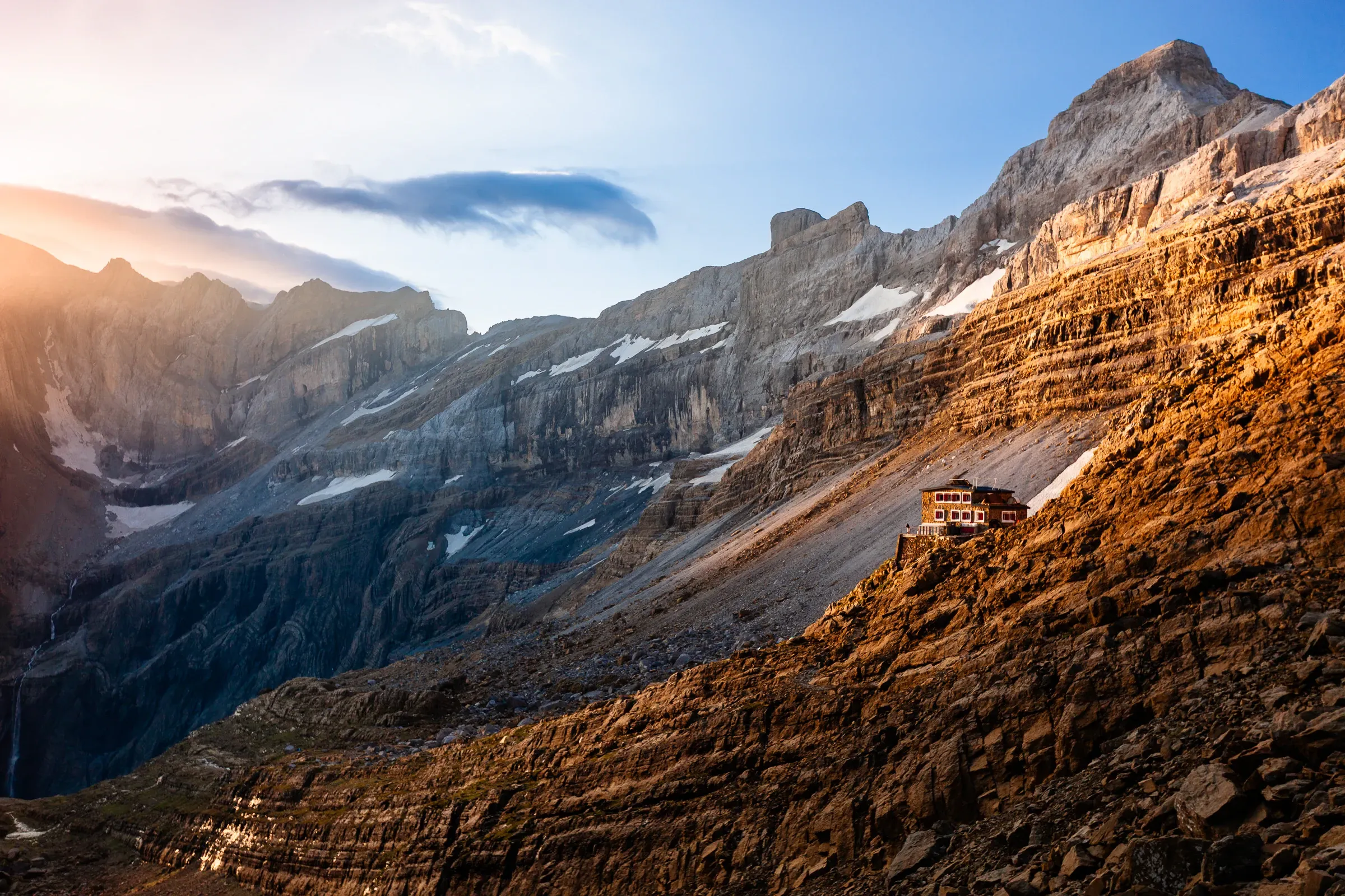Mountain refuge perched on a rocky face at golden hour, Pyrenees — dramatic cliffs rising above, warm last light on the stone walls