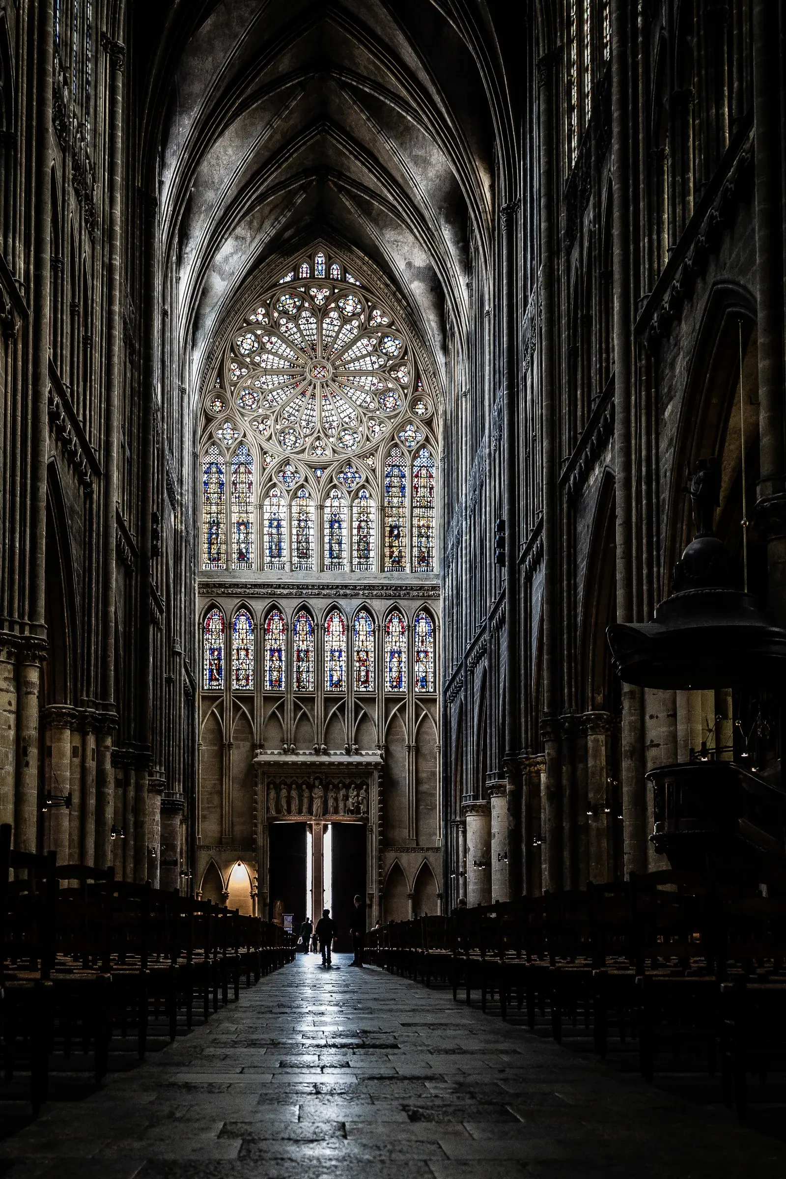 Nave of Metz Cathedral looking toward the choir, soaring Gothic arches, enormous rose window and stained glass at the far end, two figures silhouetted at the entrance