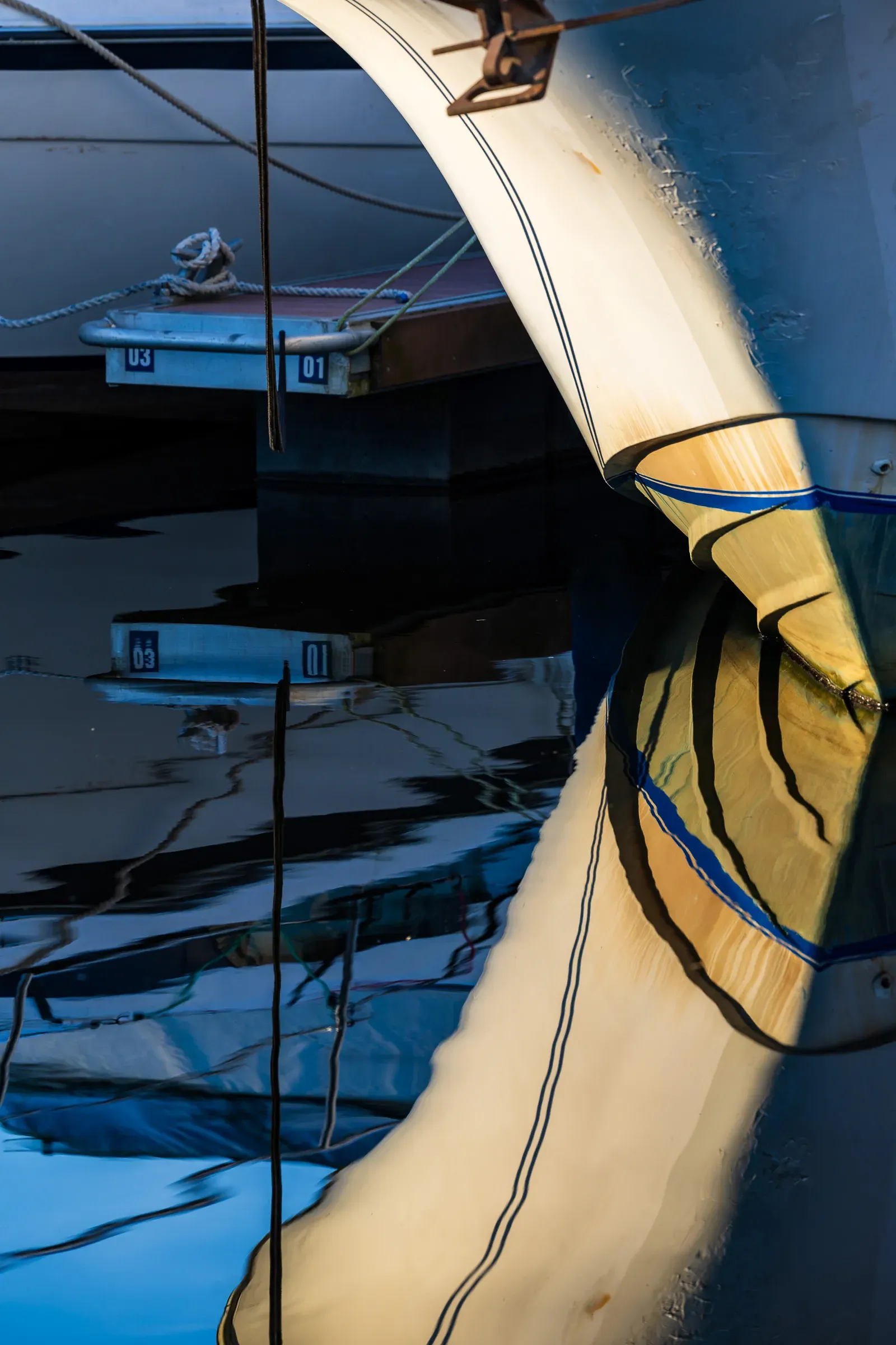 Close-up of a sailboat hull and wrapped sail with rope, numbered dock buoys reflected in dark water below