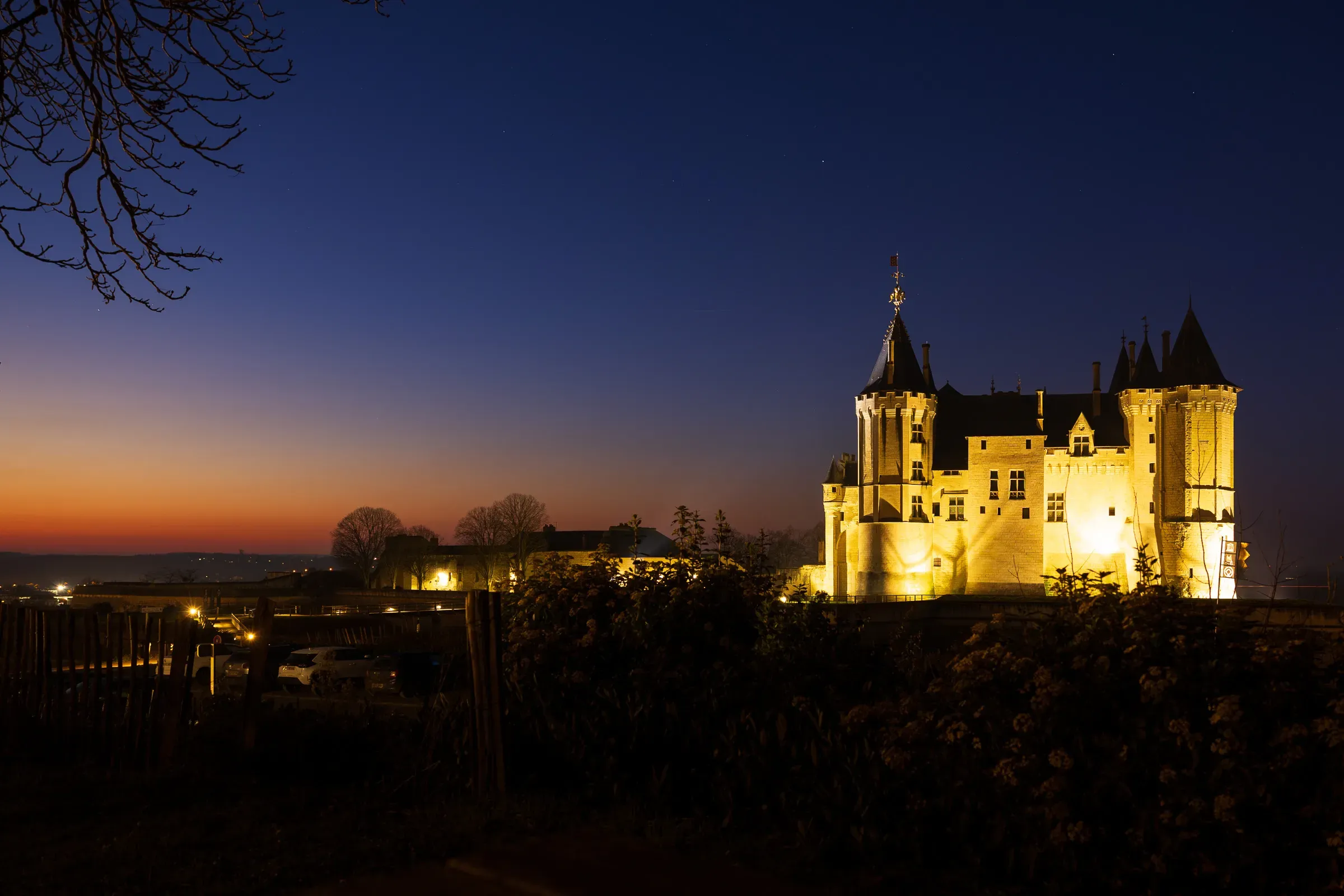 Château de Saumur floodlit at dusk, warm golden light on white stone towers against an orange-to-blue gradient sky, bare branch in the foreground, Saumur