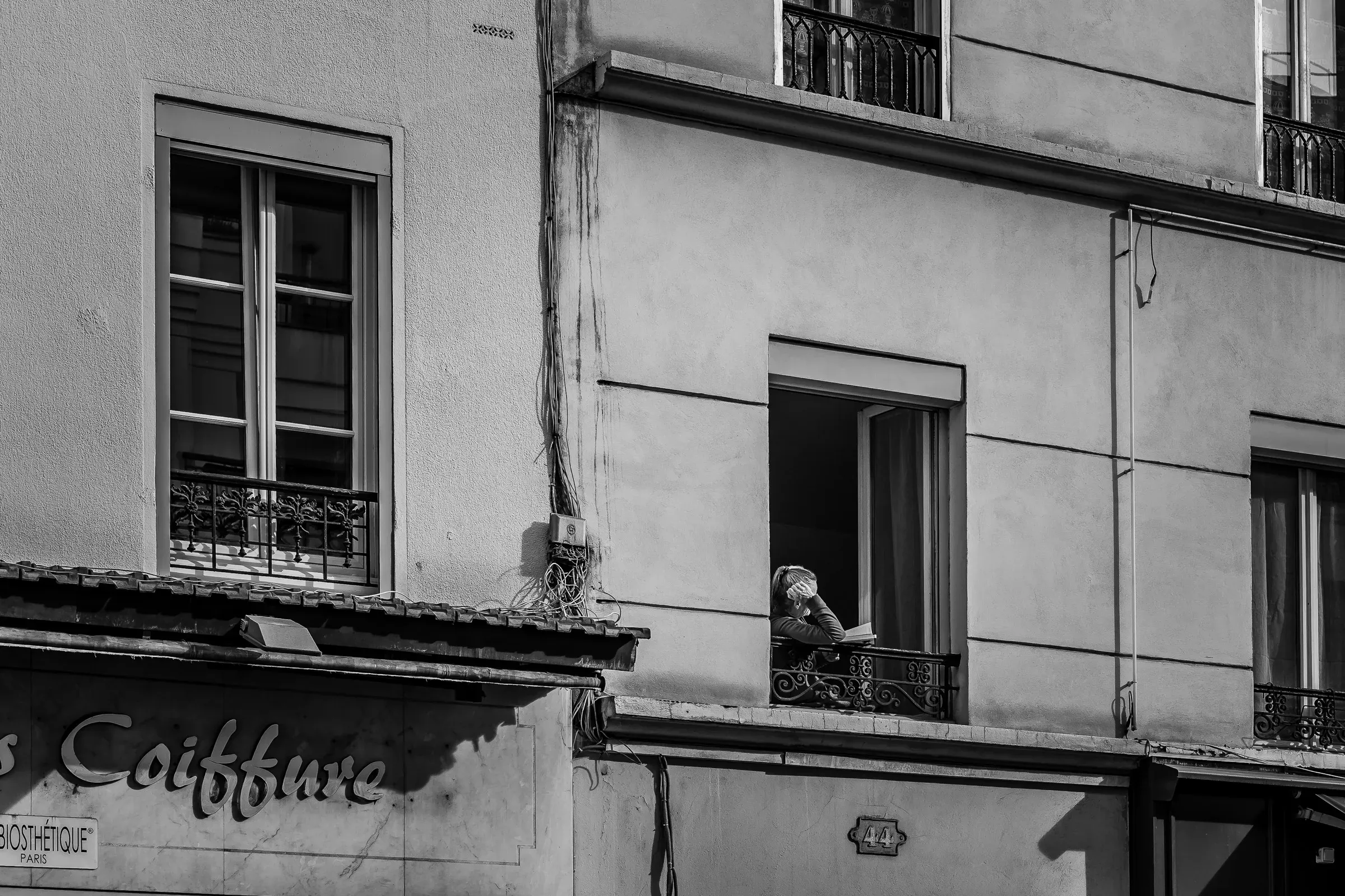 A woman reads at an open window above a hair salon sign reading 'Coiffure'
