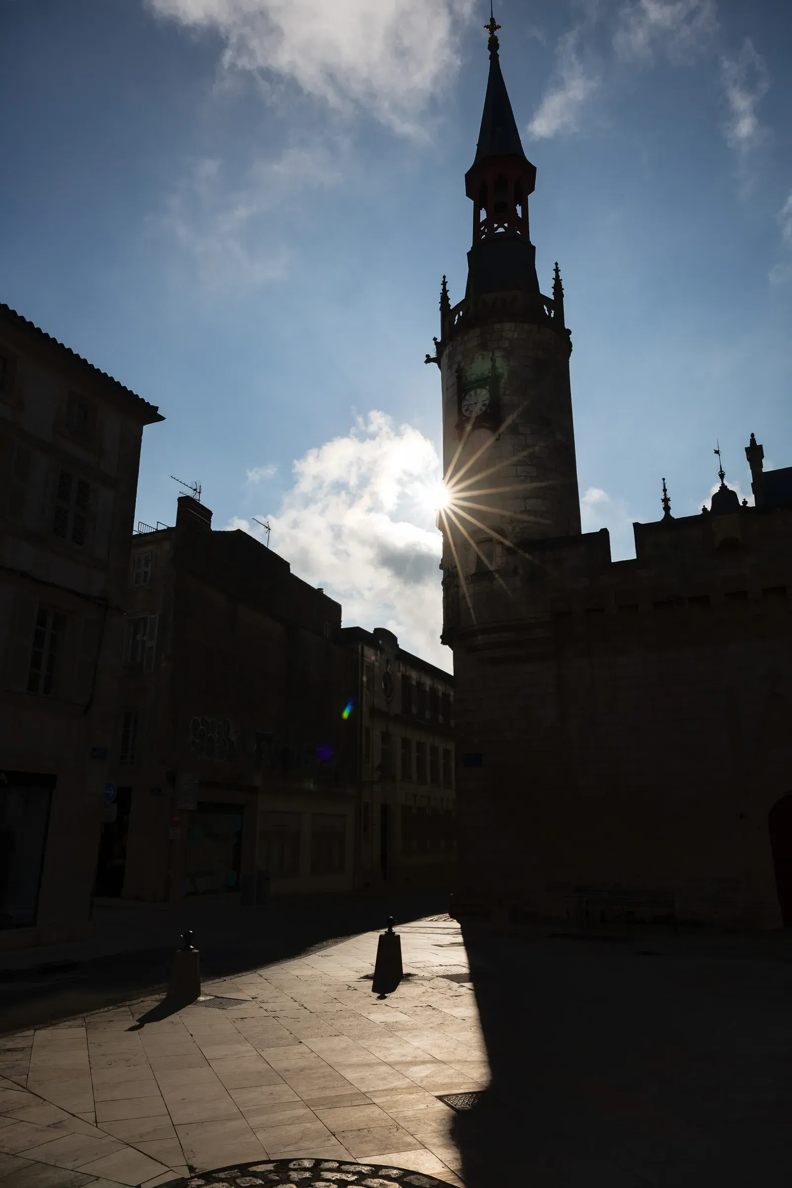 Clock tower silhouetted against a bright blue sky, sunstar bursting between the tower and a building wall, dark cobbled square with bollards in the foreground