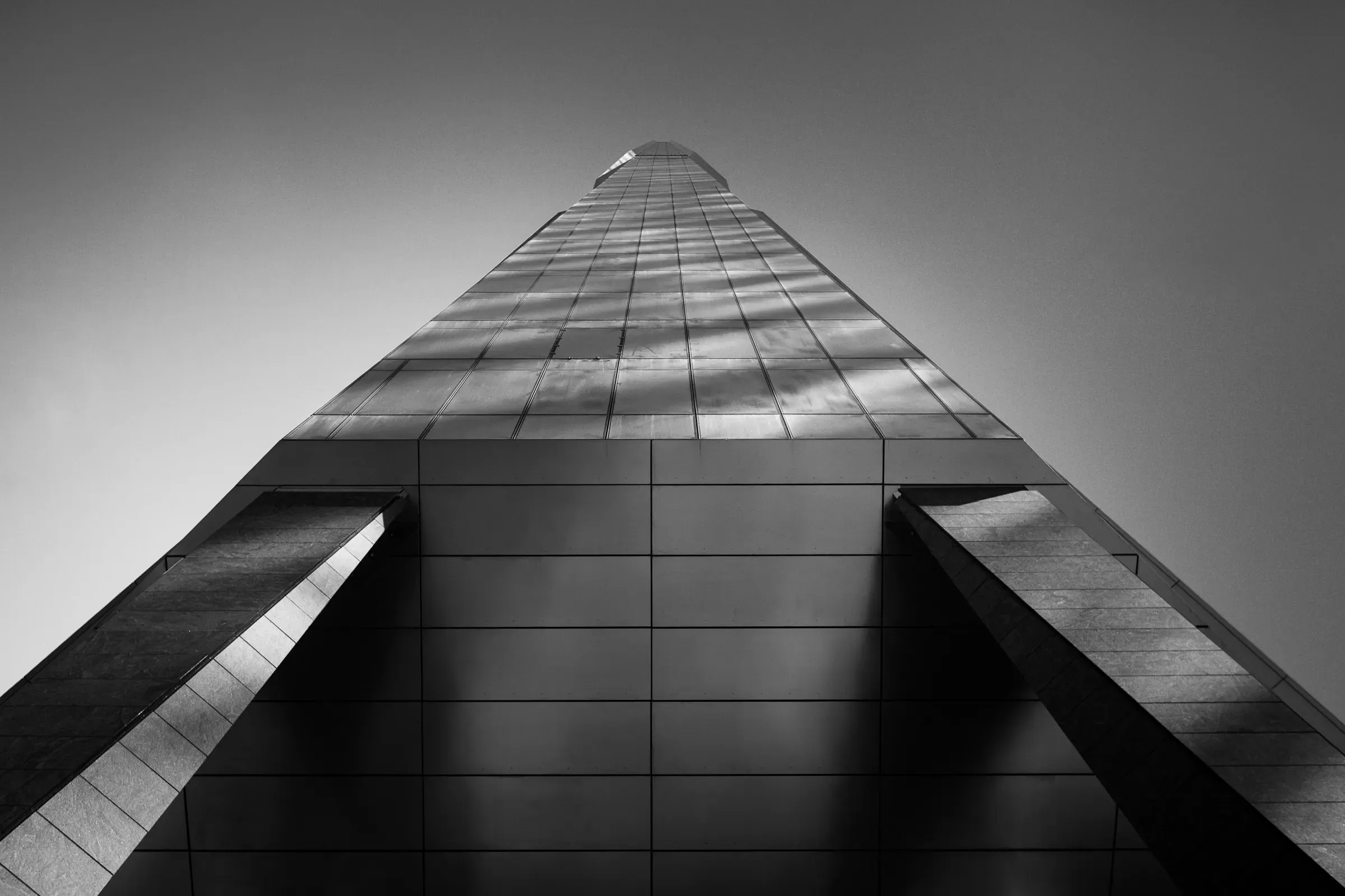 Modern glass skyscraper shot from directly below, its reflective facade receding symmetrically to a point, clouds visible in the panels