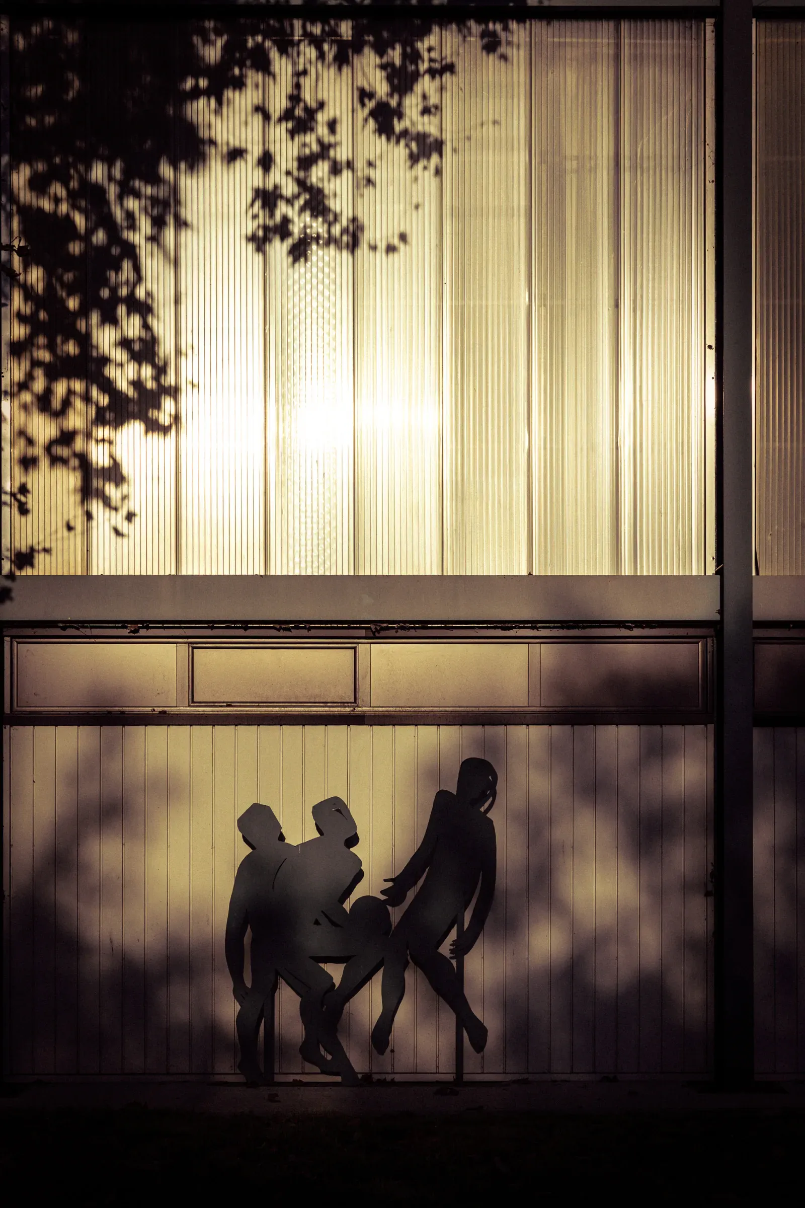 Silhouette of three figures against a glowing ribbed glass facade at night, tree branch shadows above, Bordeaux