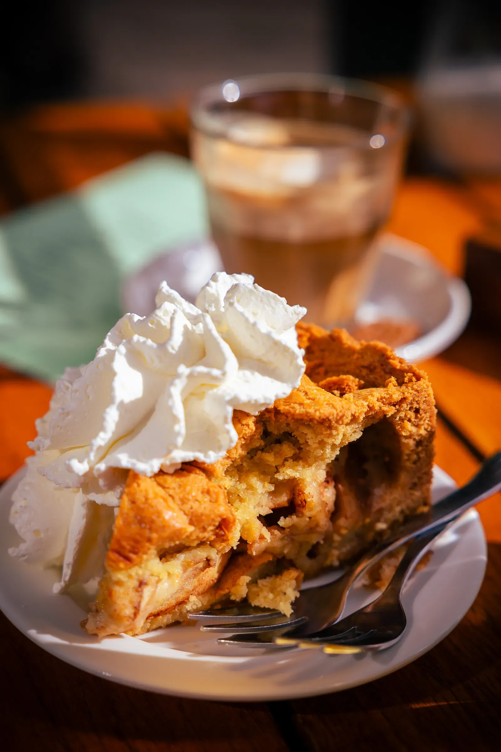 Slice of Dutch apple pie topped with whipped cream on a sunlit table, iced coffee in the background