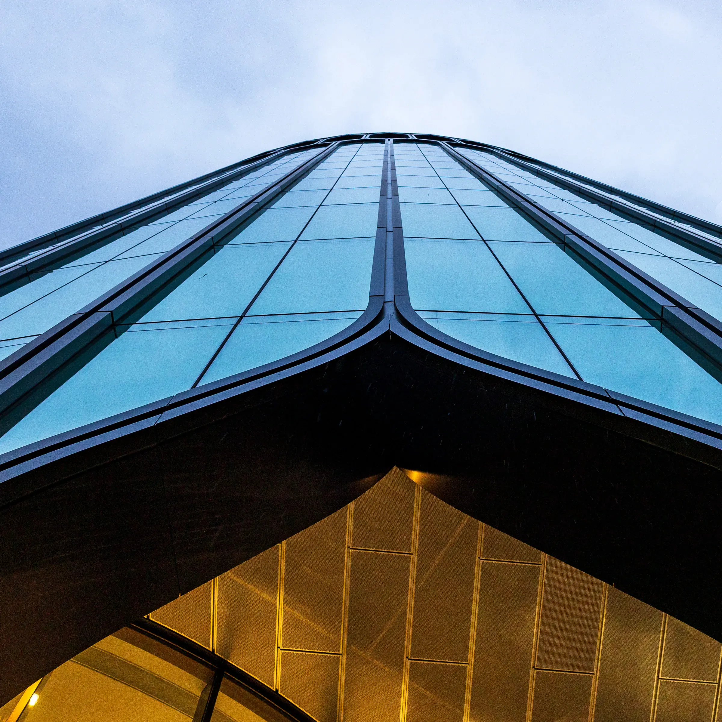 Looking up at the sharp glass corner of a Canary Wharf skyscraper, teal curtain wall tapering to a point, lit lobby visible below at dusk