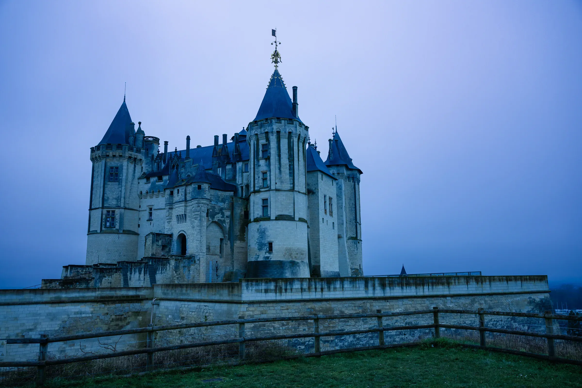 Château de Saumur at blue hour, pale stone towers under a deep blue-grey sky, wooden fence in the foreground, Saumur