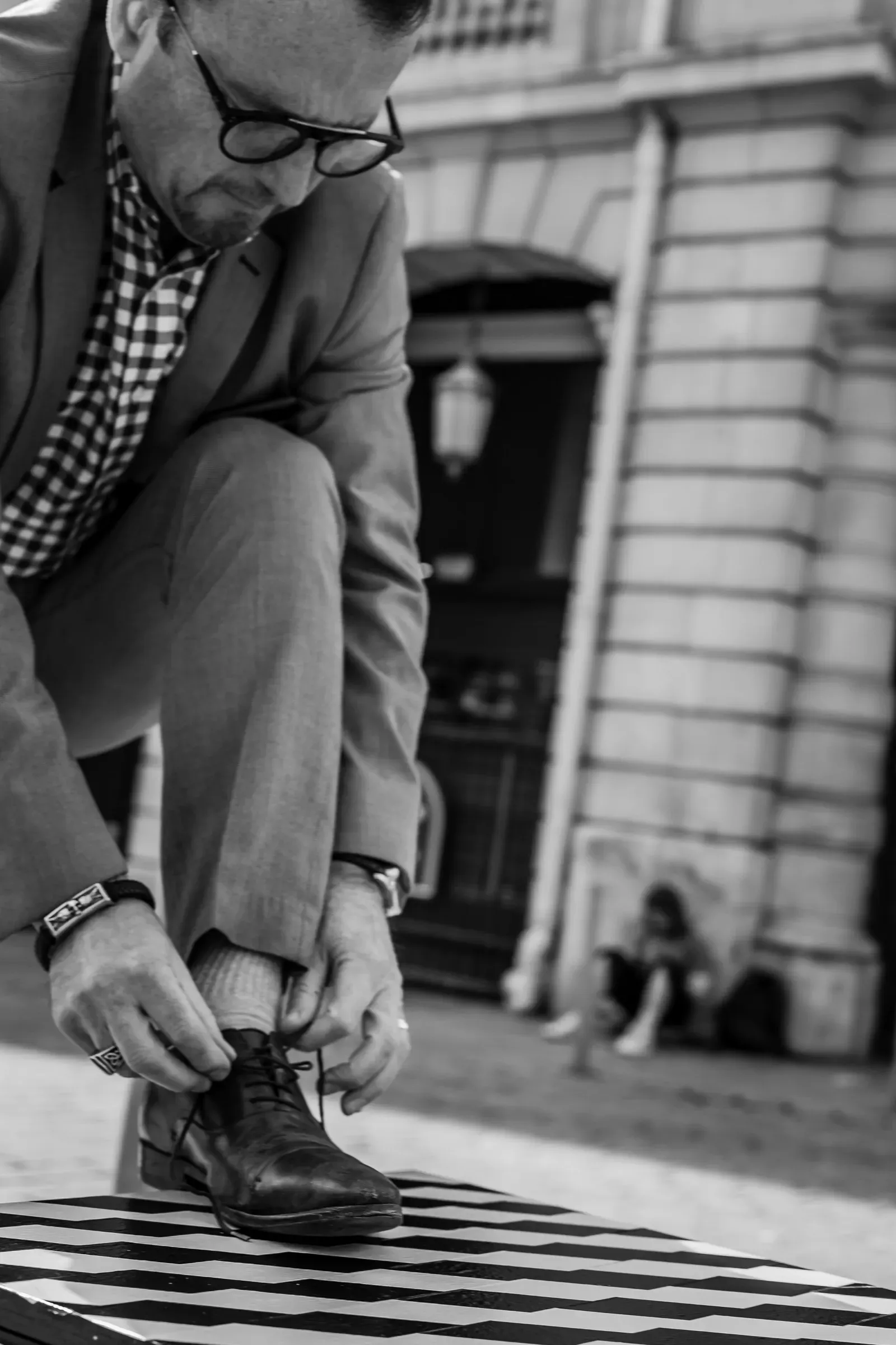 A man ties his shoelace on a café table while a blurred figure sits behind him