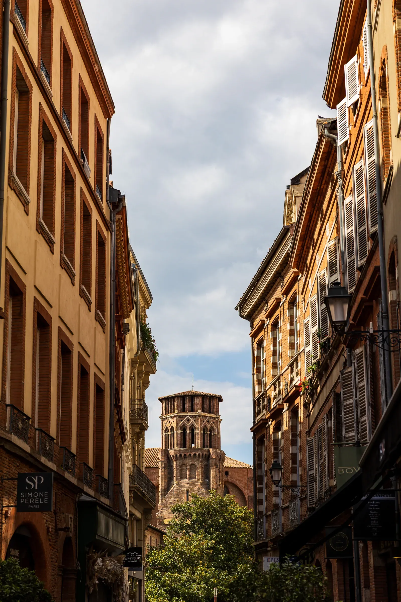 A Toulouse street looking up between tall pink brick buildings toward the ornate Romanesque tower of the Basilique Saint-Sernin, clouds above, Toulouse