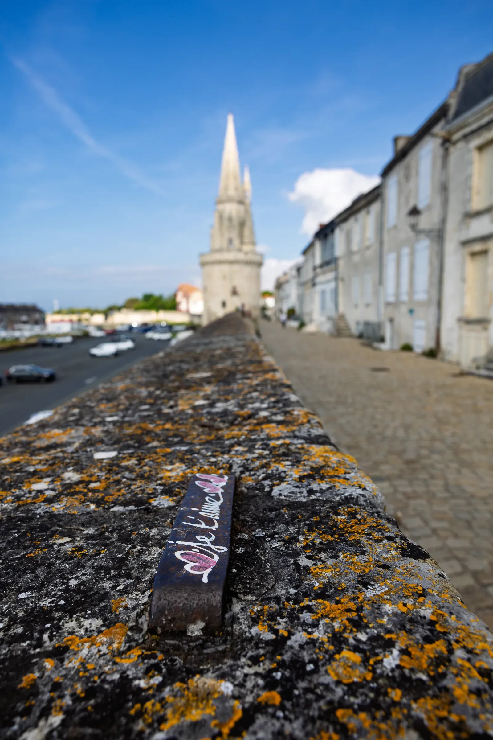 Lichen-covered stone parapet in sharp focus with a small sticker on its edge, the Lanterne tower blurred in the background along a cobbled street