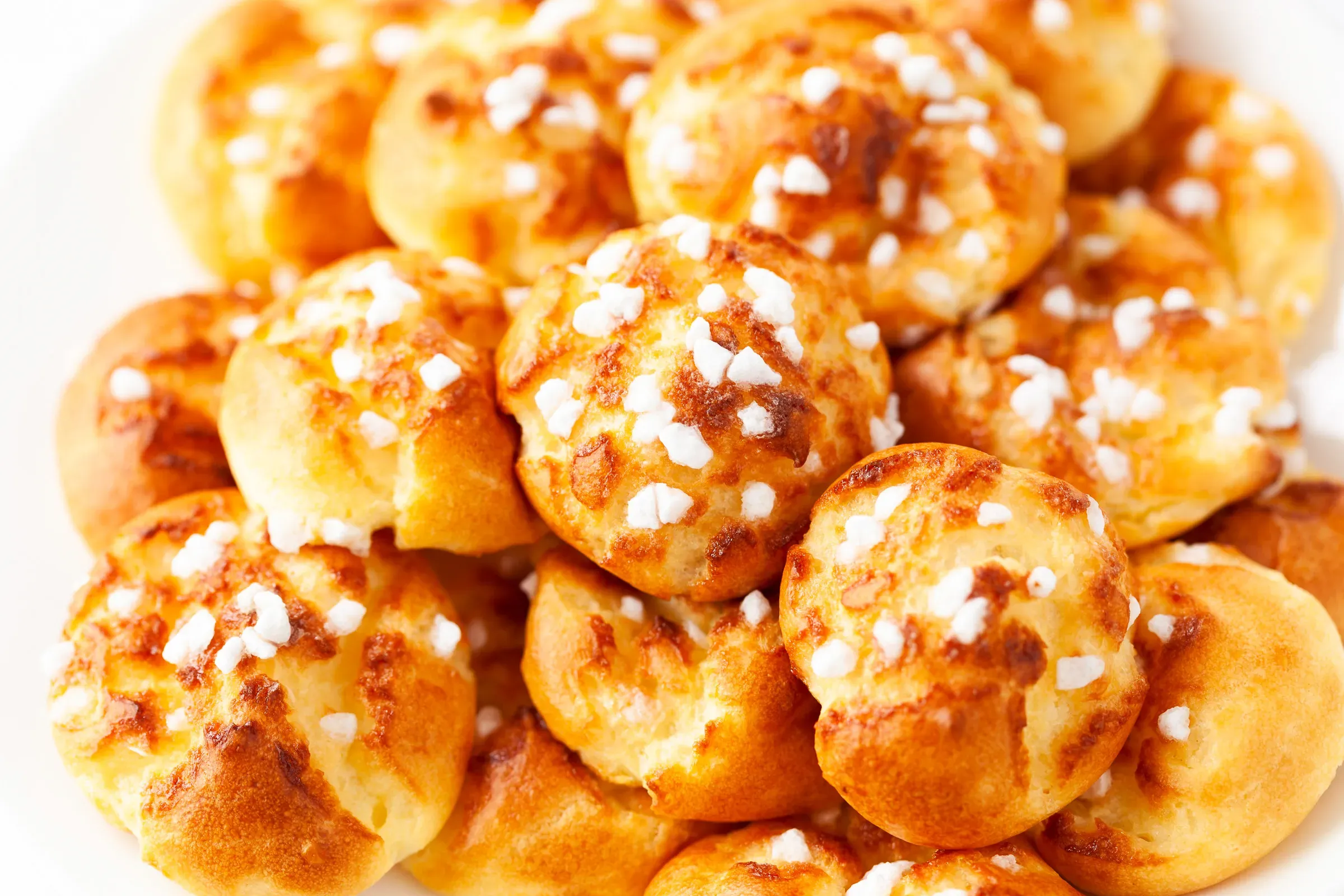 Pile of chouquettes mounded on a white plate, pearl sugar catching the light on each golden choux puff
