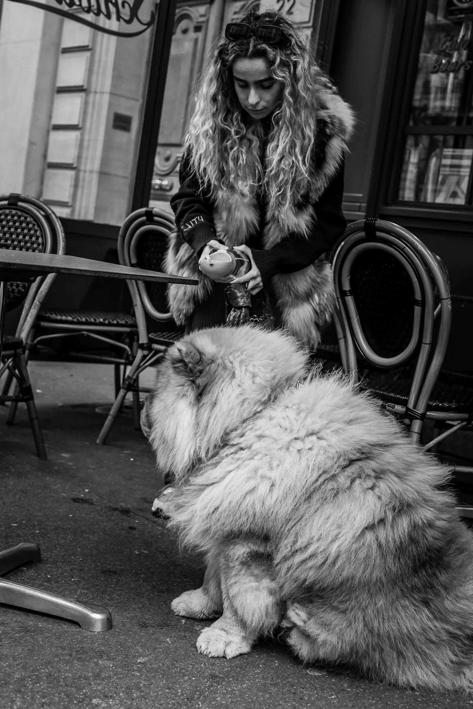 A woman in a fur-trimmed coat feeds two large fluffy dogs outside a café
