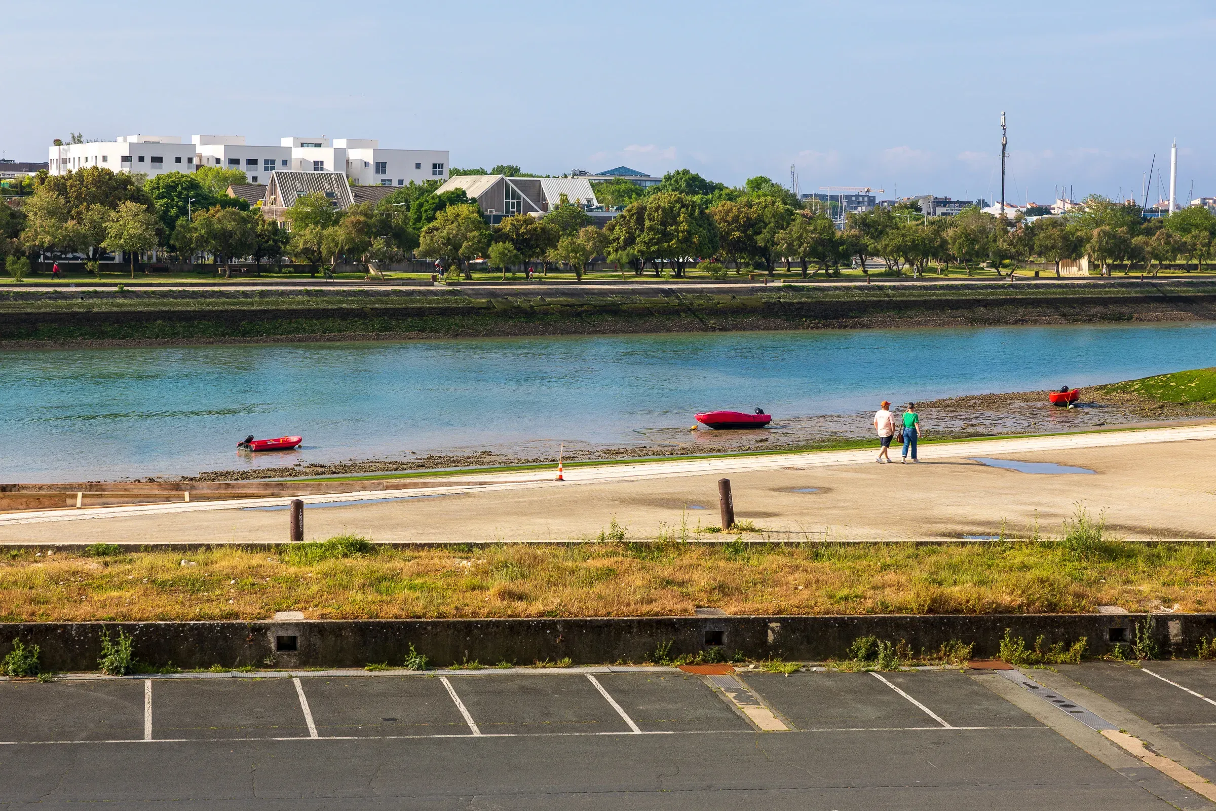 Two red kayaks and a couple walking on a sandy strip beside calm turquoise water, modern buildings and trees across the inlet, empty car park in the foreground