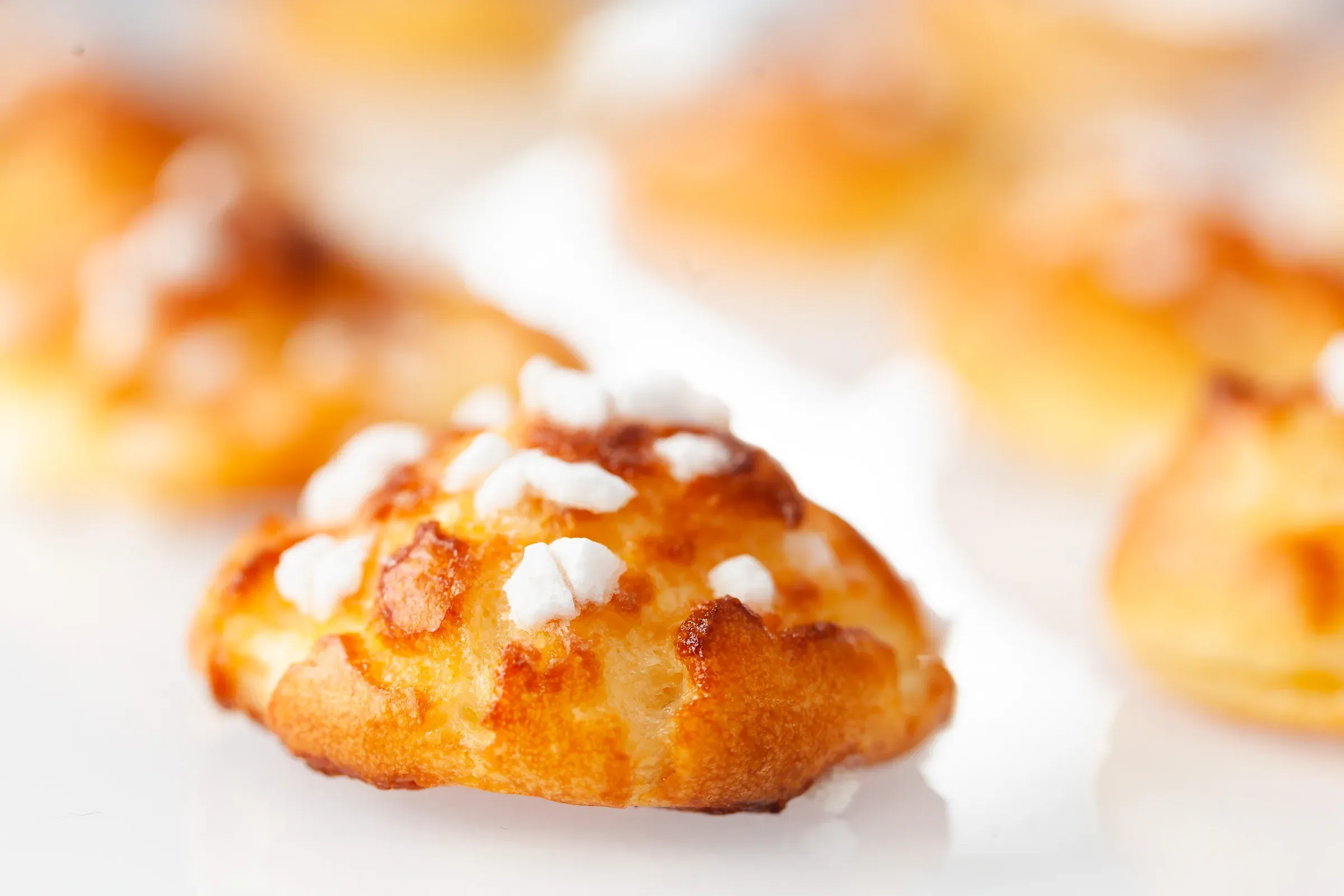 Single chouquette in sharp focus, pearl sugar crystals on the domed top, other puffs soft behind on a white surface