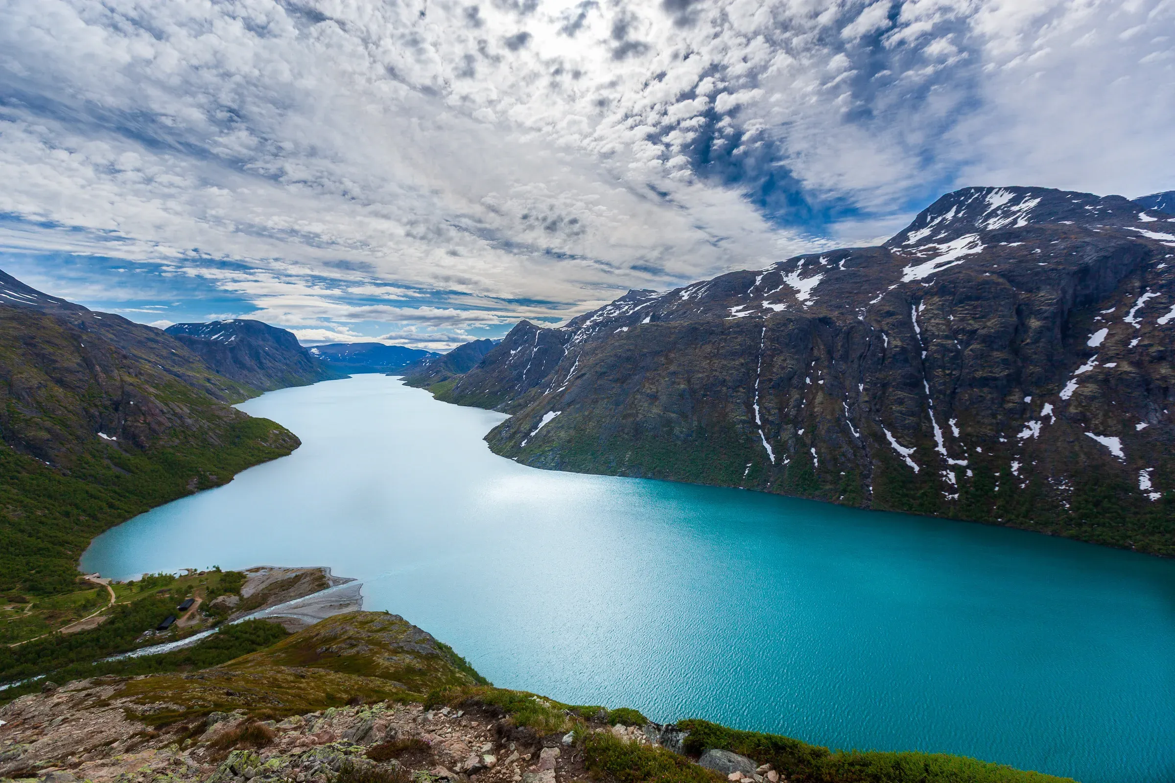 Lake Gjende from the Besseggen ridge, Jotunheimen — turquoise water curving between steep snow-patched mountains, dramatic cloud ceiling