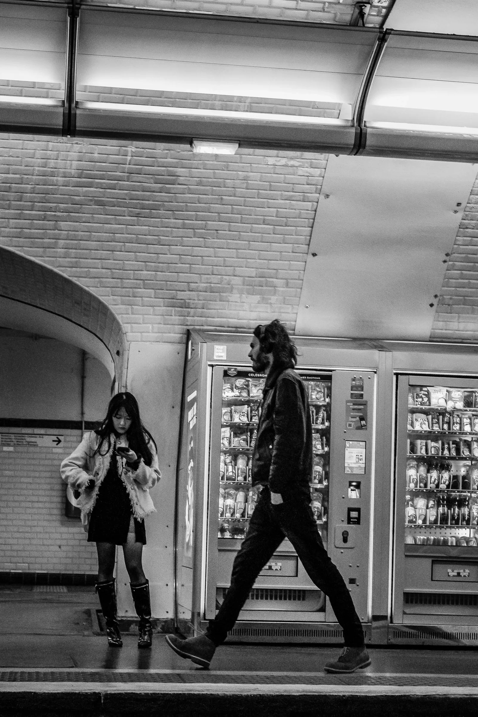 A woman checks her phone on a metro platform as a man walks past a row of vending machines