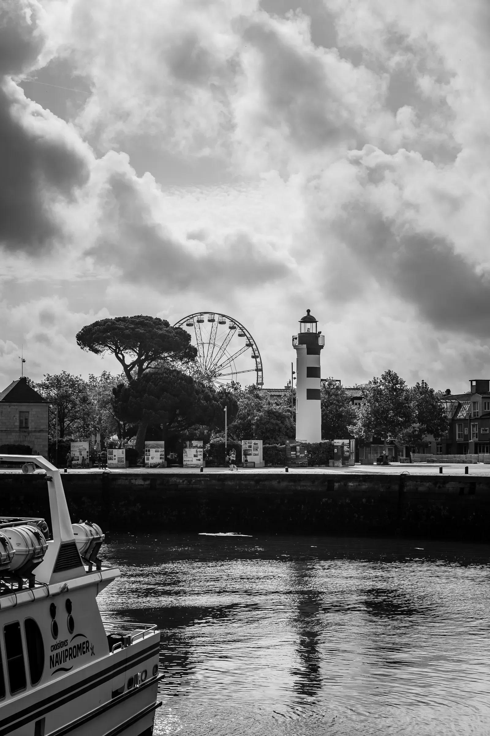 Black and white — harbour view with a boat bow in the foreground, La Rochelle's lighthouse and Ferris wheel in the mid-distance, dramatic cumulus clouds above