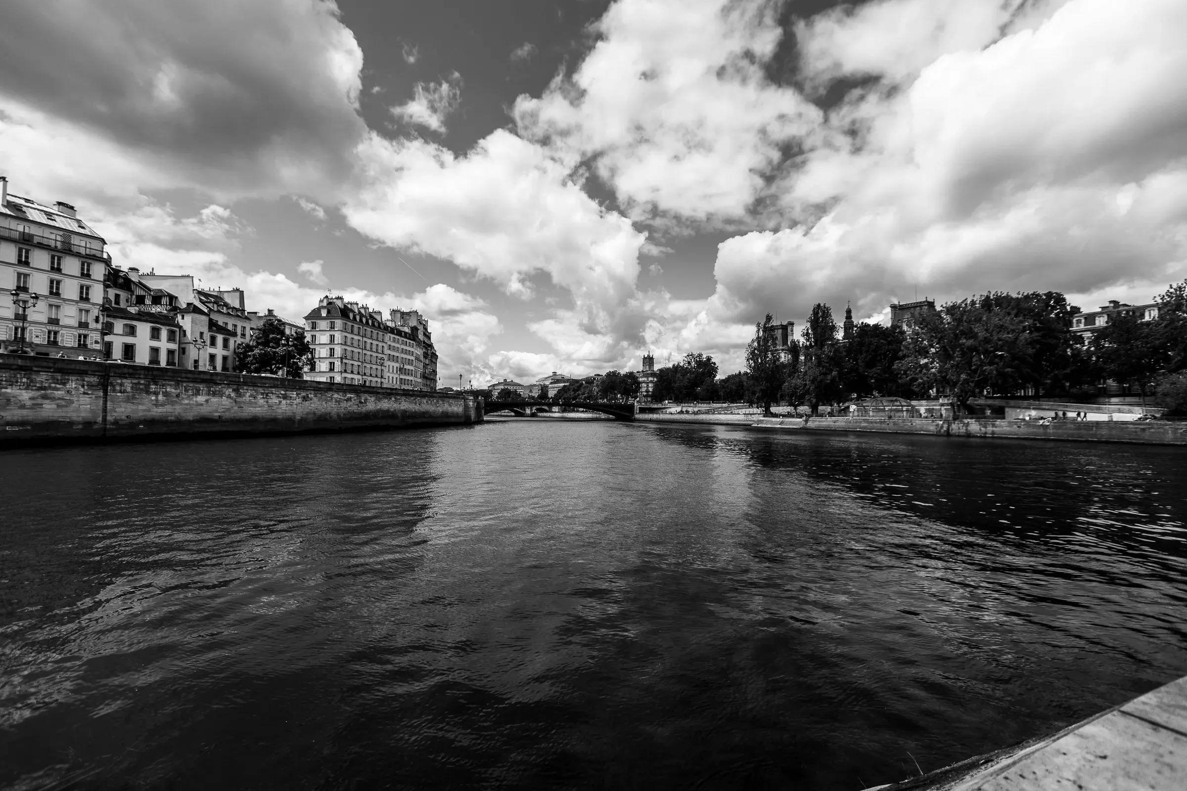 River Seine in Paris looking west, Haussmann apartment blocks lining the left bank, a bridge in the middle distance, voluminous clouds above