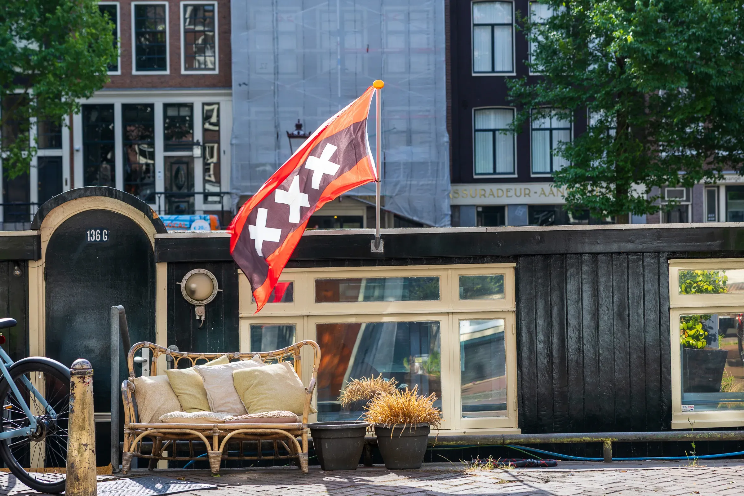 Amsterdam flag with three white crosses flying from a houseboat deck, rattan sofa and canal houses behind