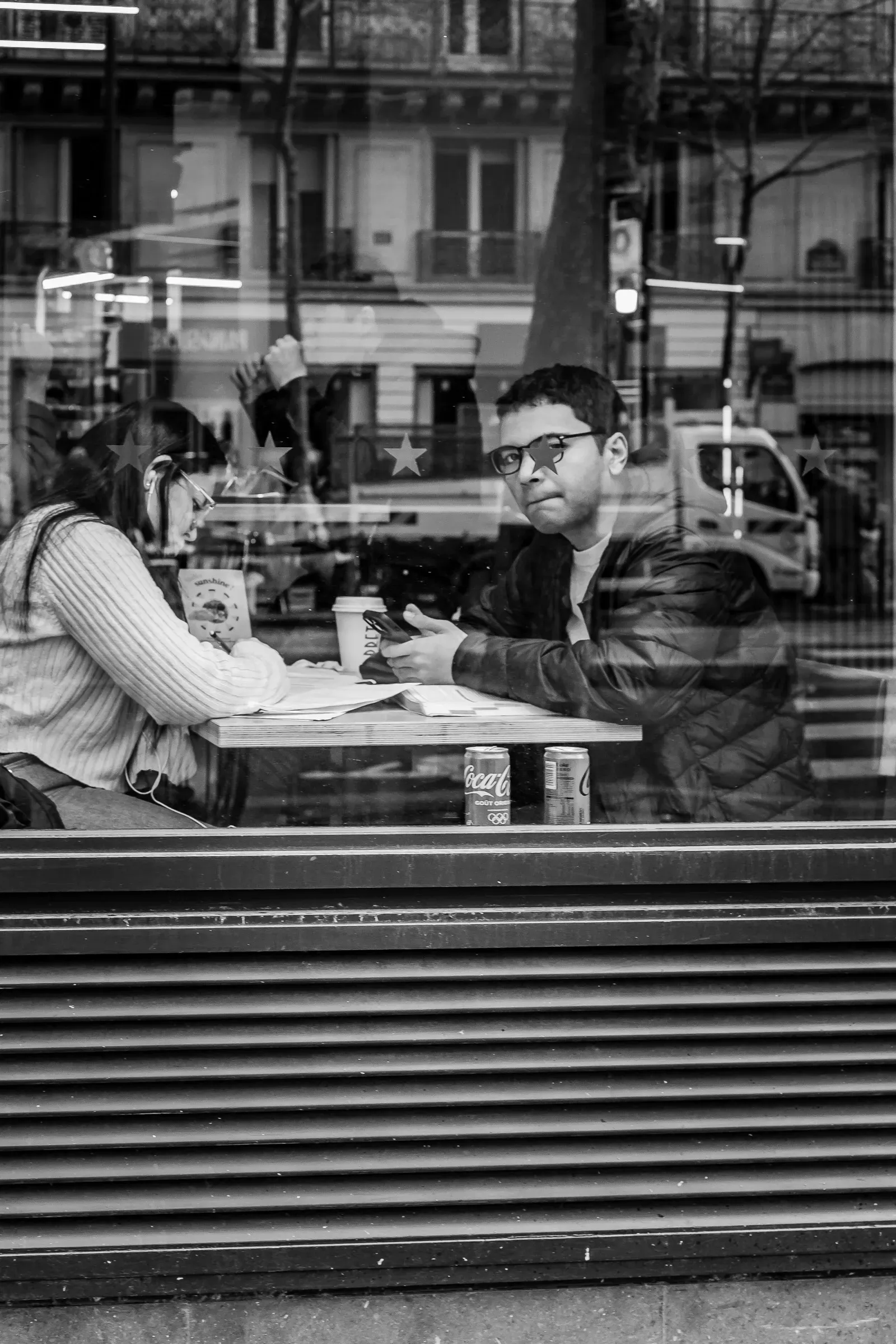 Two people at a café table seen through a reflective window, one looking up from his phone toward the camera